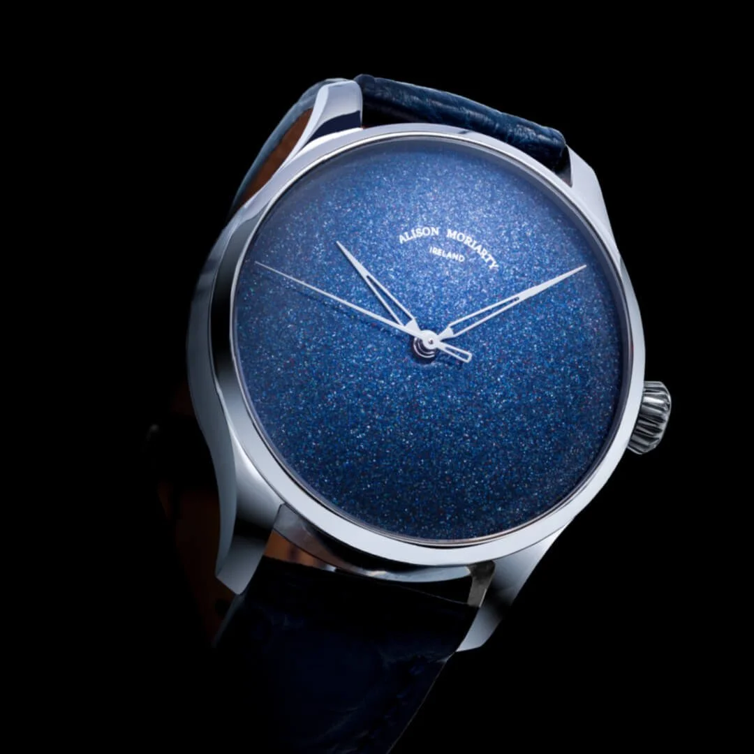 Close-up of a luxury watch with a blue, glittery face, silver case, and black leather strap, displaying the brand name Alison Moriarty Ireland.
