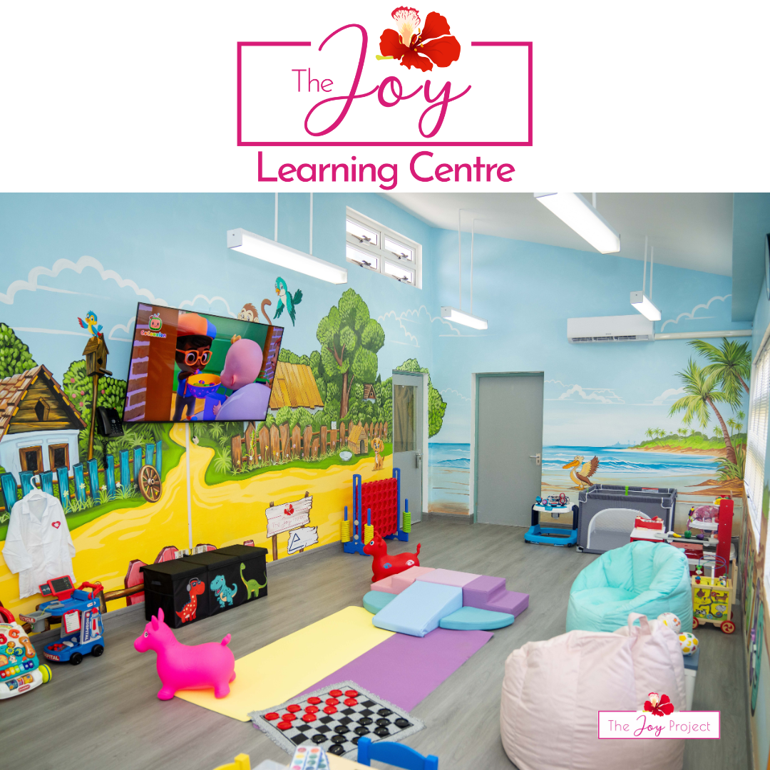 Colorful indoor children's learning and play area with wall murals of a tropical beach scene, toys, play mats, and a TV screen, part of The Joy Learning Centre.