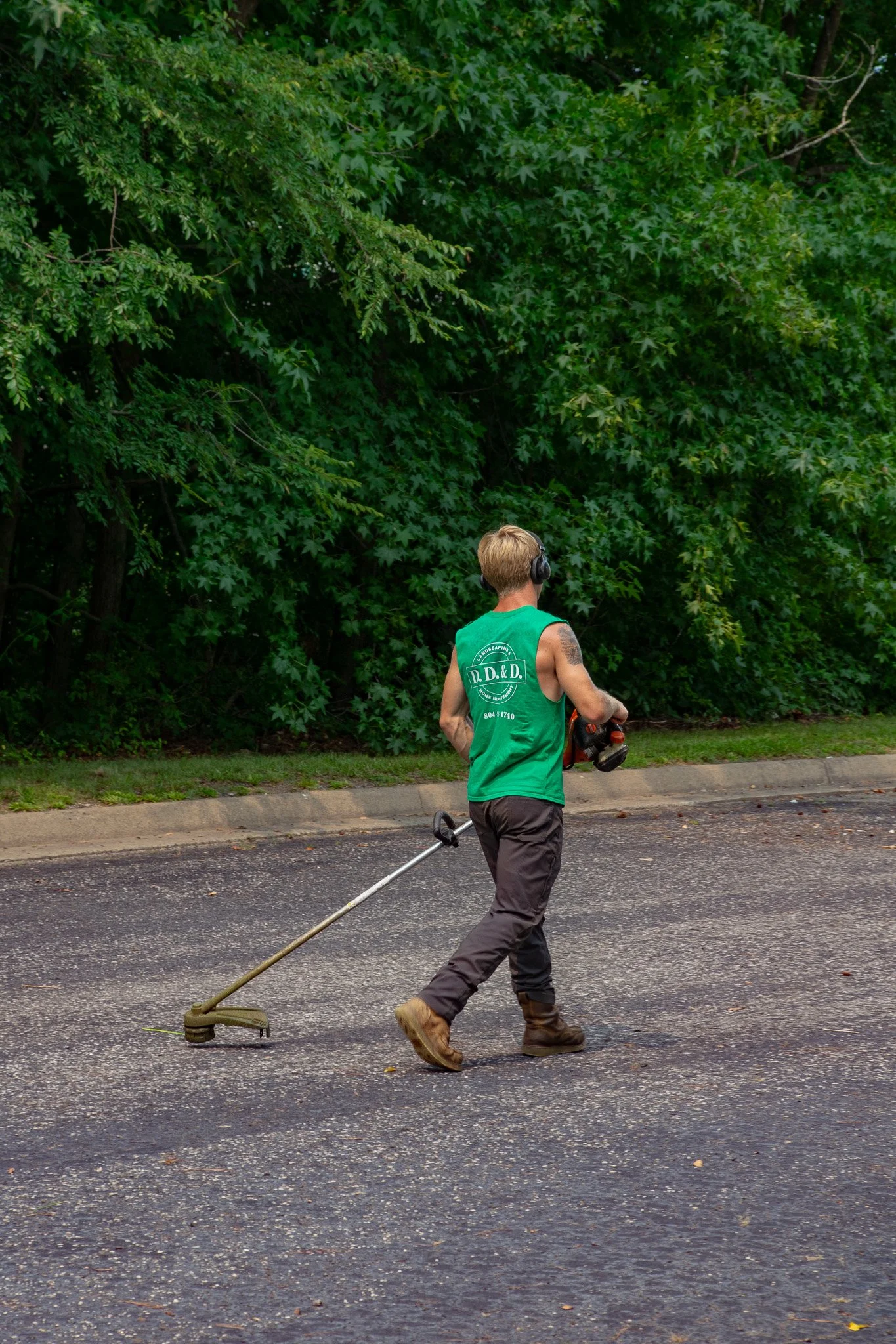A man with blonde hair wearing a green shirt, black pants, and brown boots is walking on a paved street, holding a grass trimmer and wearing black headphones. There is a dense leafy green tree in the background.