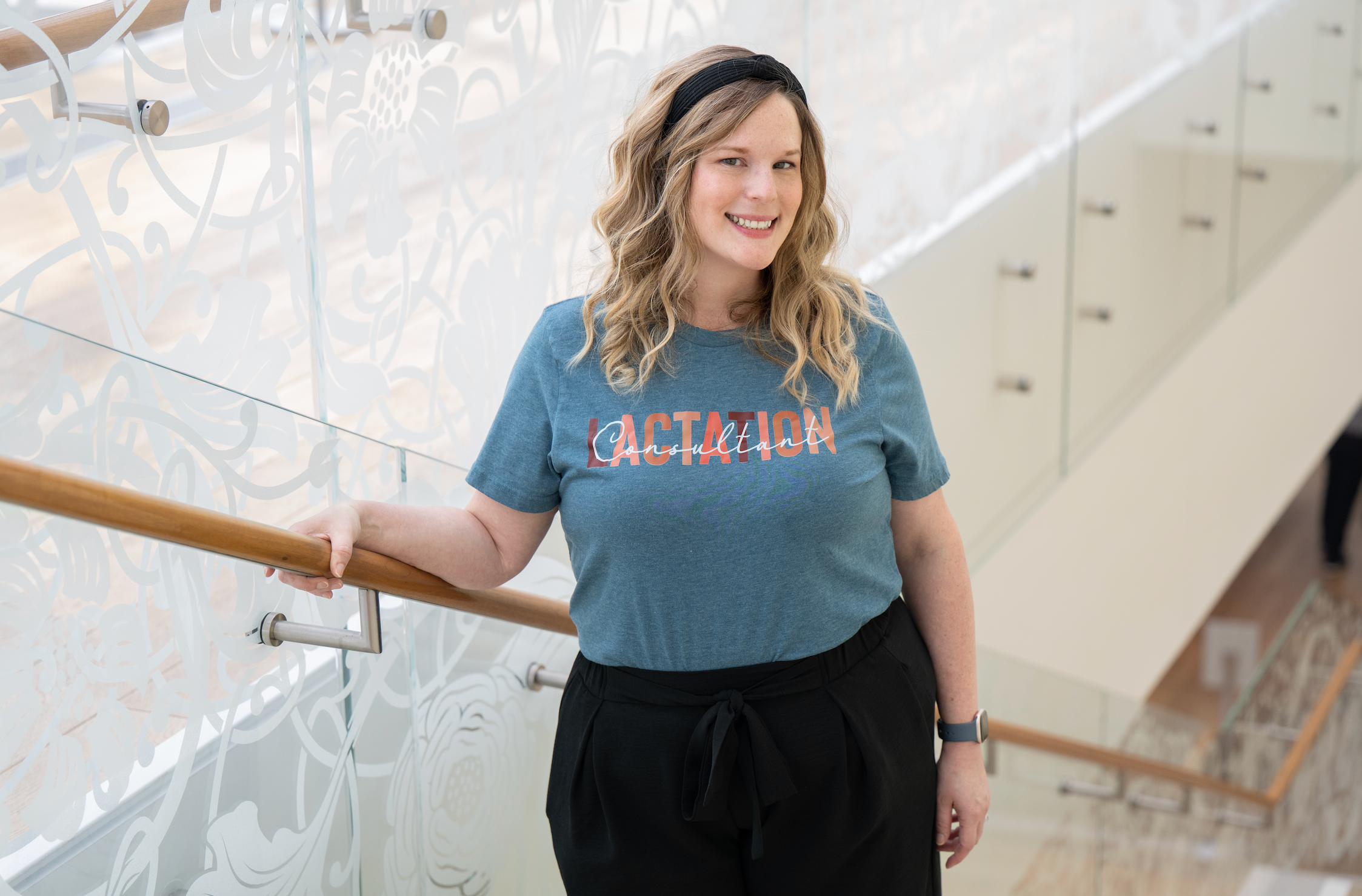 A smiling woman with wavy blonde hair wearing a blue T-shirt that says 'Lactation' standing indoors next to a glass wall with decorative patterns and a staircase with a wooden railing.