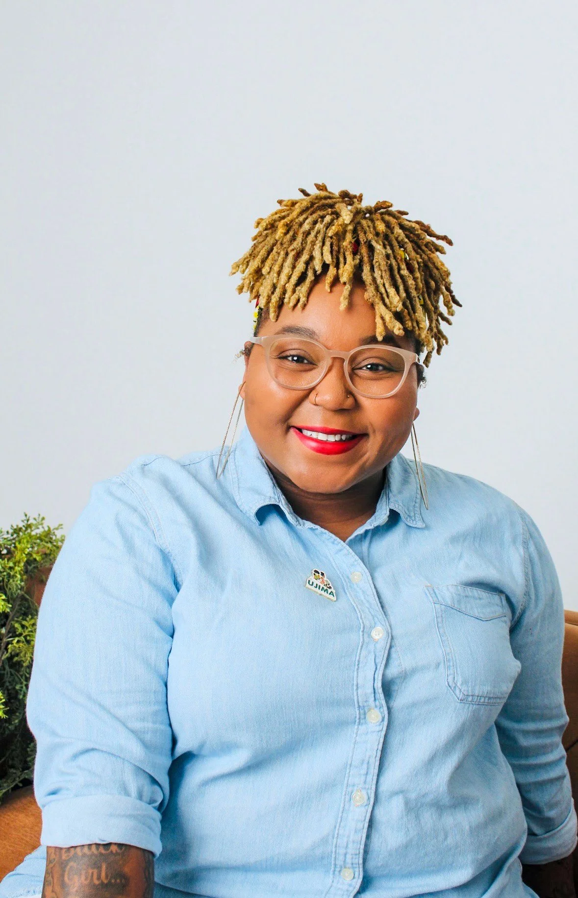 A smiling woman with glasses and dreadlocks, wearing a light blue button-up shirt with a pin, sitting in front of a light-colored wall.