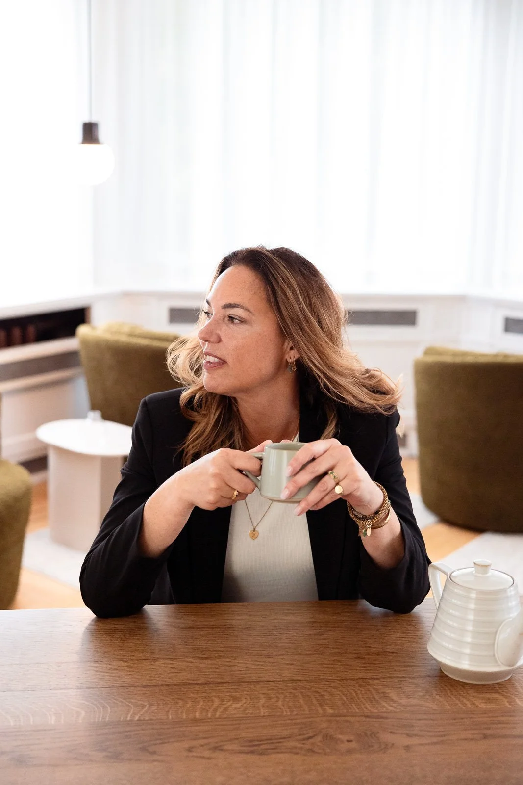 Vrouw met bruin haar in zwart jasje drinkt koffie uit een lichtgroen kopje in een modern binnenhuis met groene stoelen en houten tafel.