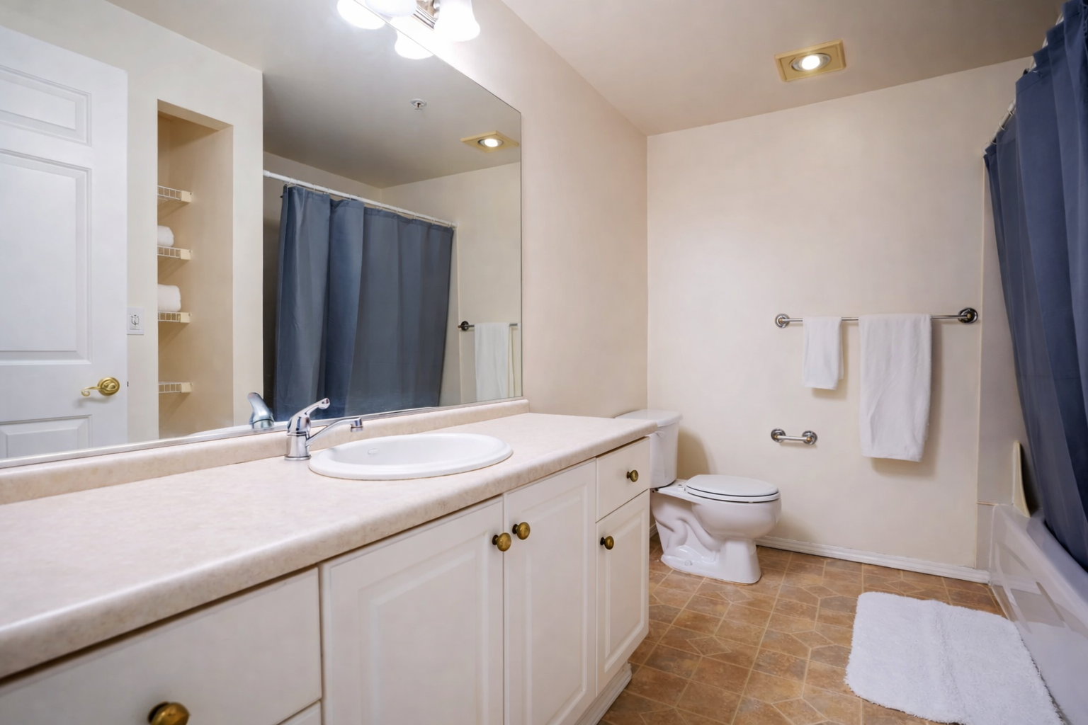 Bathroom with a large mirror, beige countertop, white cabinetry, toilet, blue shower curtain, white towels on a metal rack, beige tiled floor, and recessed lighting.