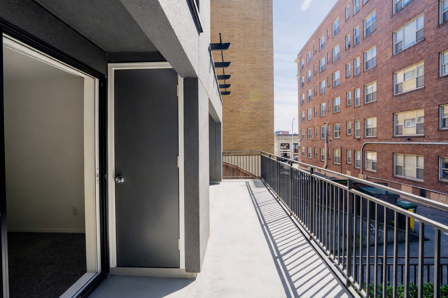 Apartment balcony with open door leading inside, black railing, and neighboring brick buildings with multiple windows.