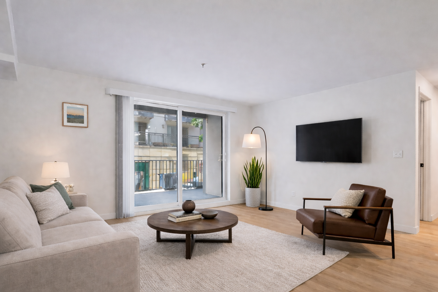 Modern living room with white walls, sliding glass door leading to balcony, beige sofa with pillows, wood coffee table, brown armchair with pillow, floor lamp, television on wall, potted plant, framed picture, and hardwood floor.