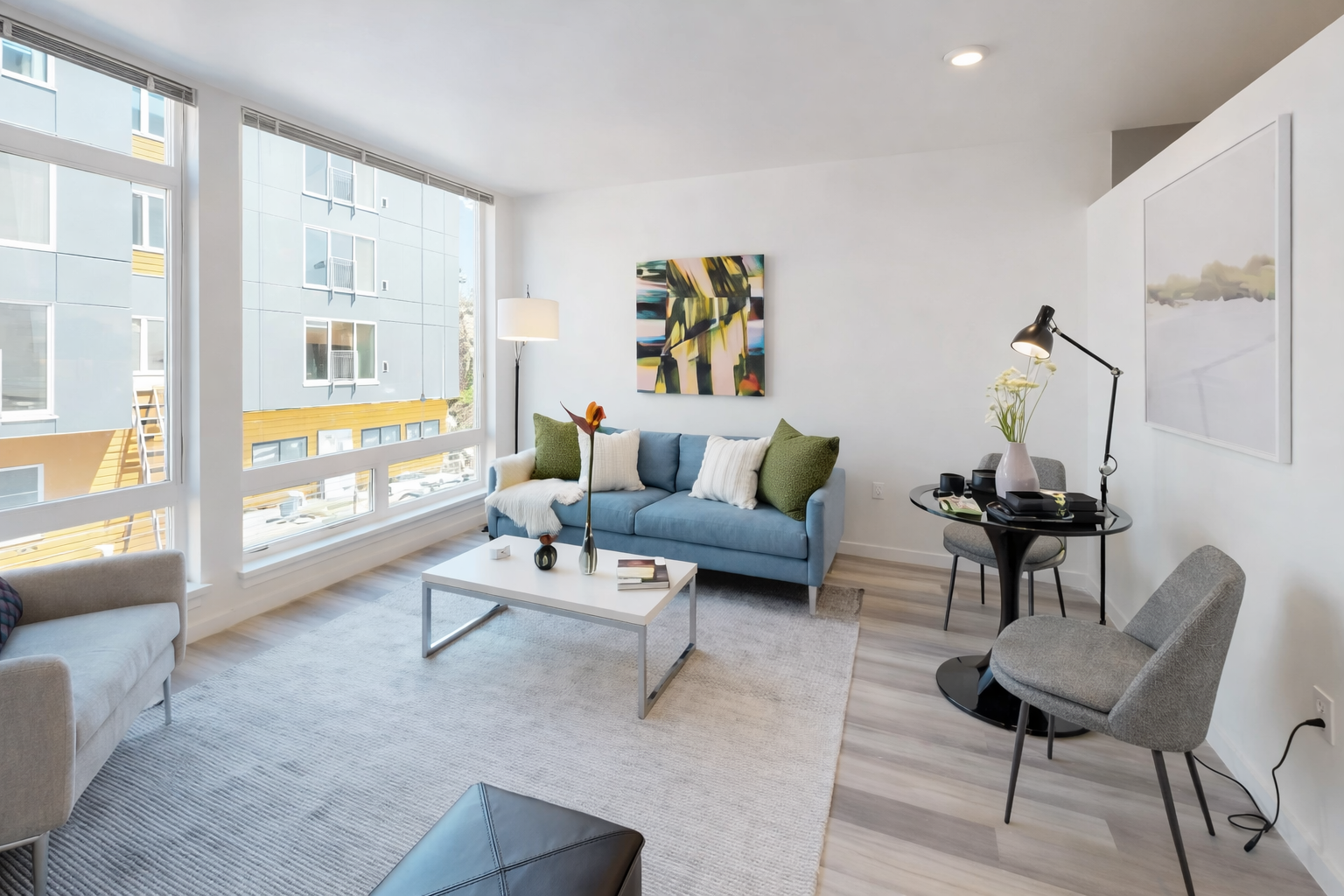 Modern living room with large windows, blue sofa with cushions, white coffee table with decorative vases, wall art, armchair, and small black accent table with a lamp and decorative items.