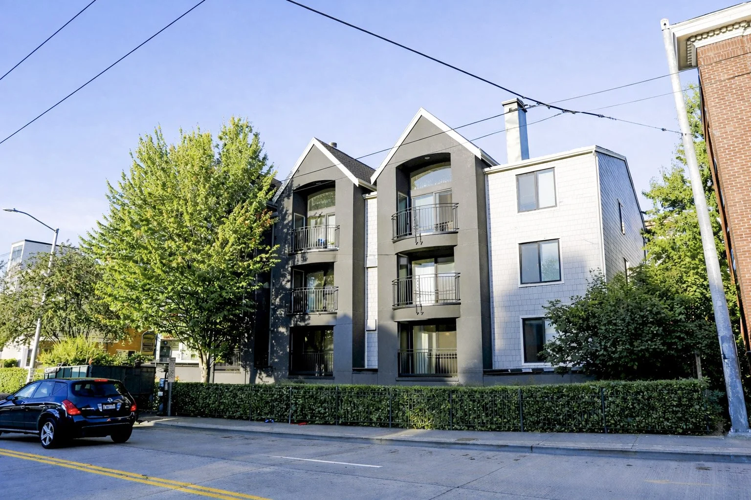 Multi-story residential building with balconies, surrounded by trees and greenery, parked car on the street, overhead utility lines, clear blue sky.
