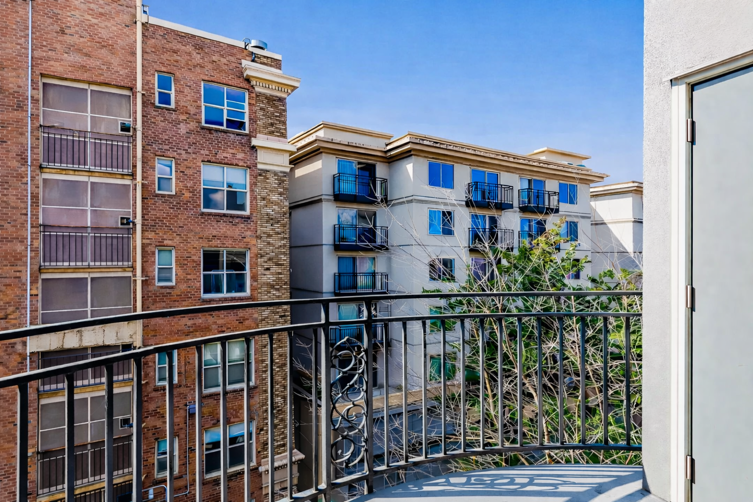 View from a balcony showing neighboring apartment buildings with windows and balconies, some with black metal railings, under a clear blue sky.