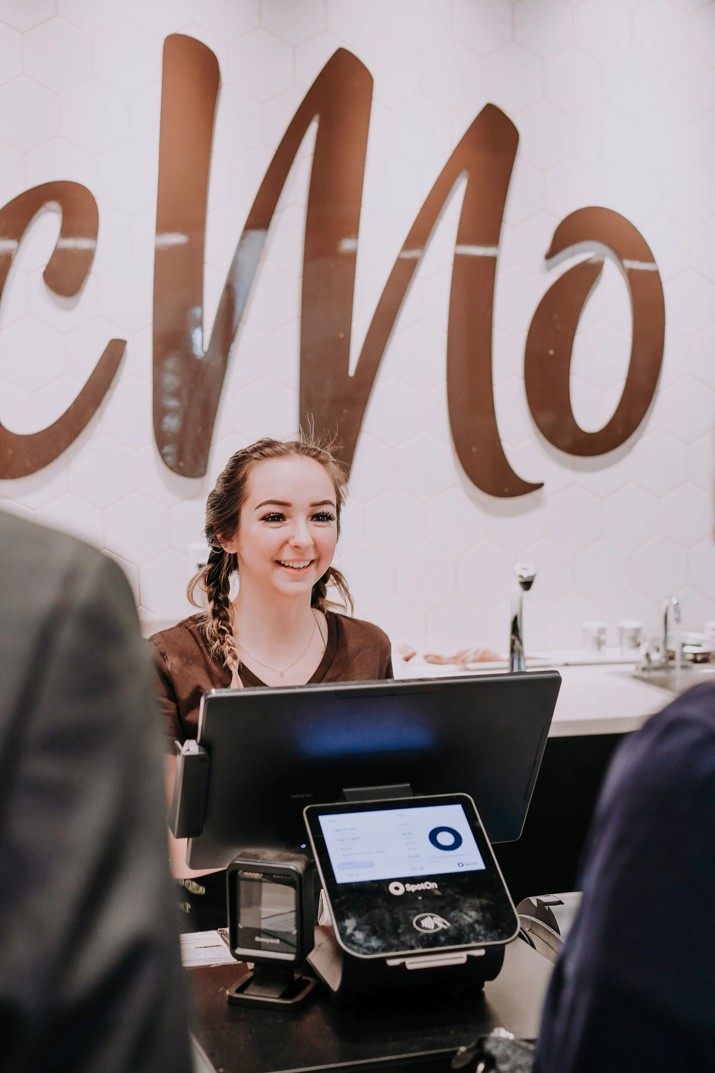 A woman at a register smiling while interacting with customers at a store or restaurant, with a large decorative sign that says 'CVS' in the background.