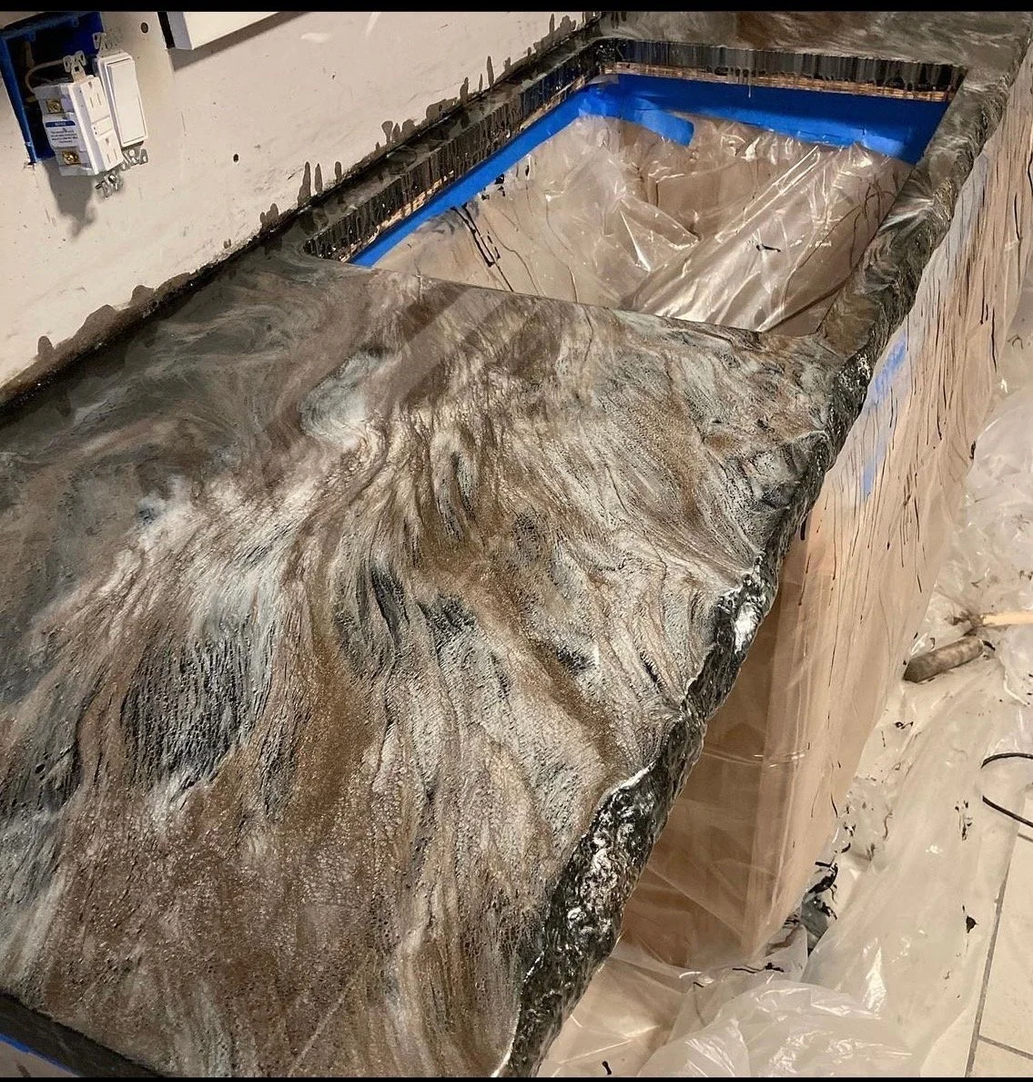 Close-up of a marble or granite countertop with a glossy, swirling pattern in shades of brown, black, and white, under construction with a protective lining inside the empty sink area.