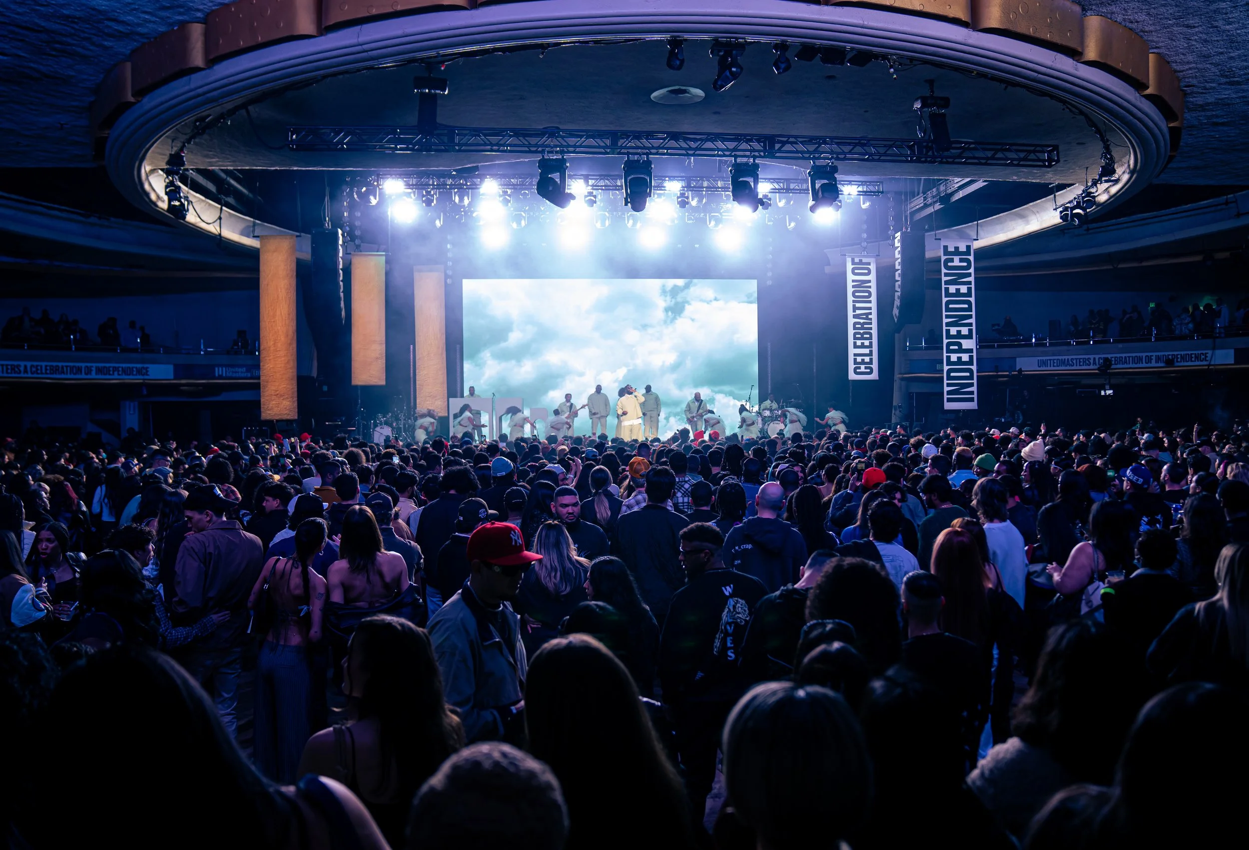 Large indoor concert with stage, bright lights, and a large crowd of people, some wearing hats, attending a celebration event.
