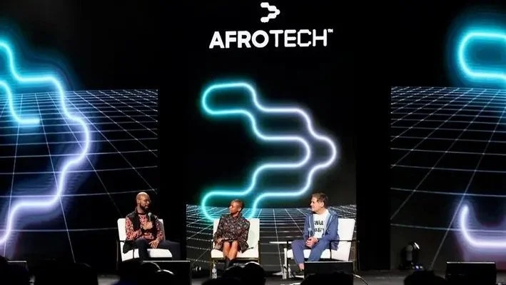 Three people seated on stage during a panel discussion at AfroTech conference, with large digital screen behind them displaying AfroTech logo and abstract neon designs.