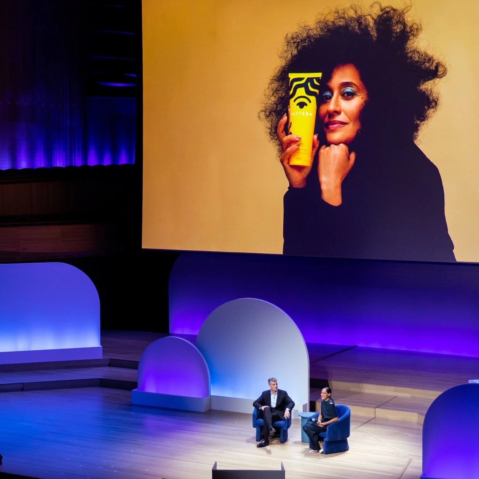 A stage with two people seated in blue chairs, a large screen showing a woman's face holding a yellow product, and decorative blue and white arches in the background. Purple lighting illuminates parts of the stage.