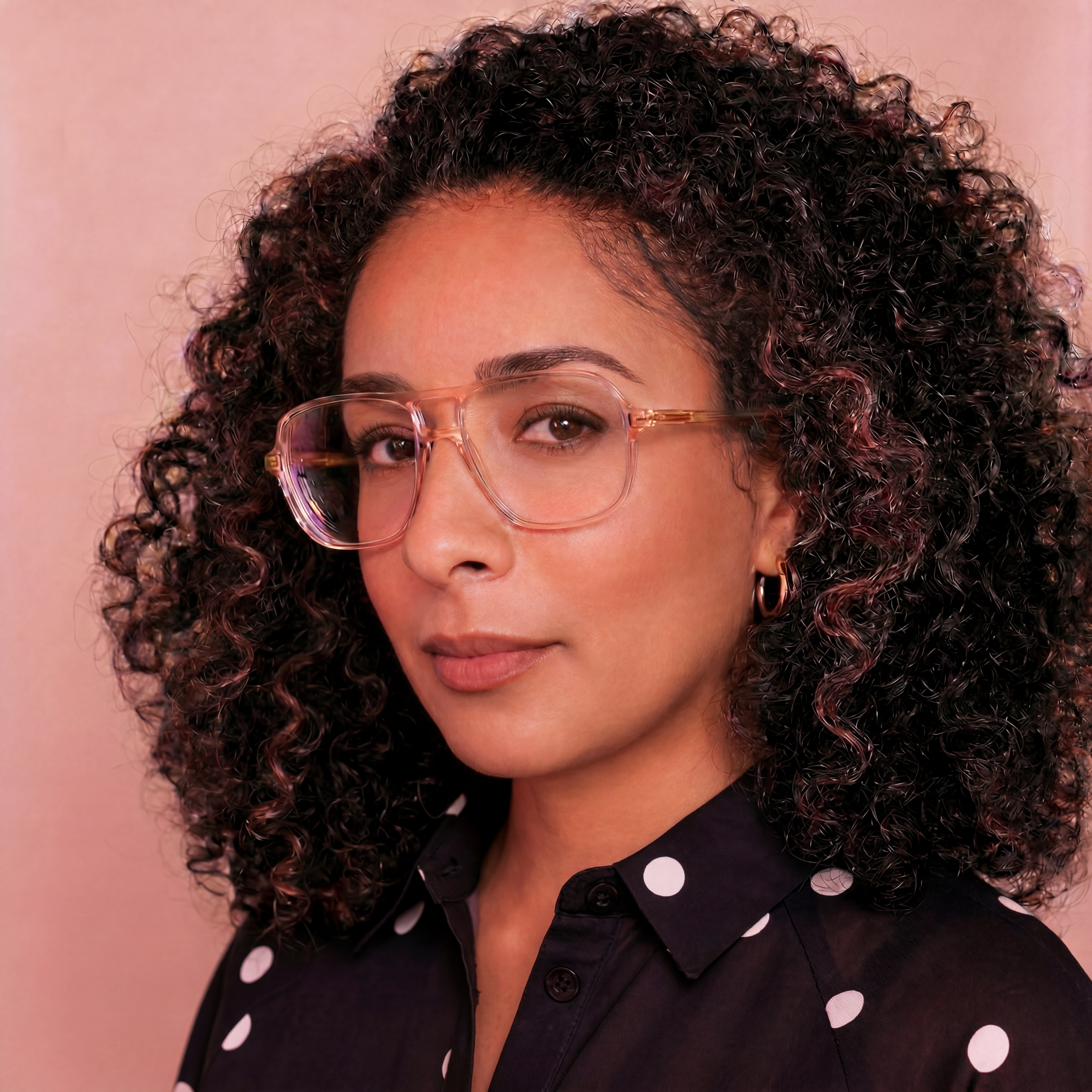 A young woman with curly hair wearing clear glasses and a black polka dot shirt against a pink background.