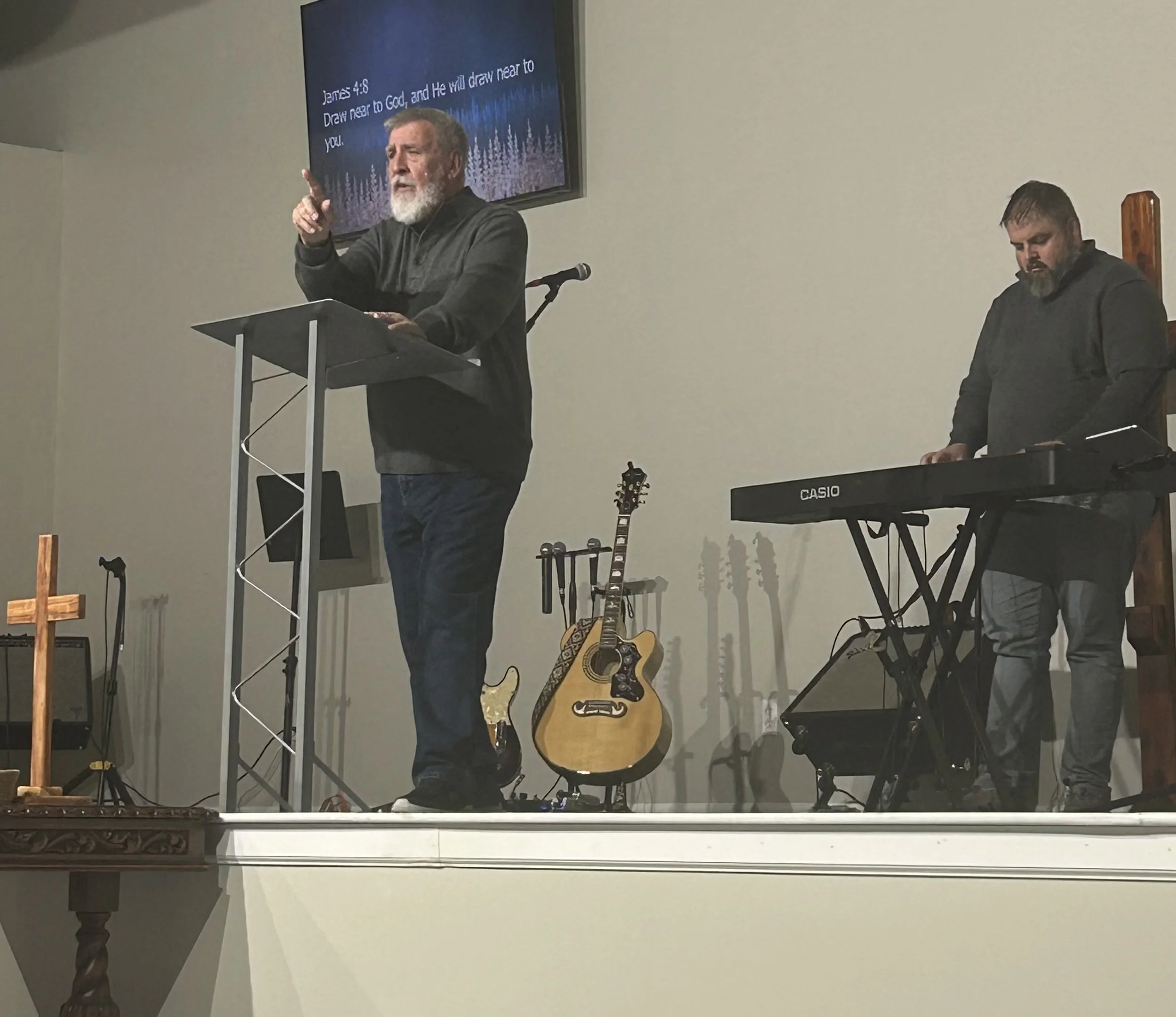 Pastor speaking at a podium with a microphone, accompanied by a musician playing a keyboard on the right, on a stage with guitars in the background, and a large screen displaying scripture behind them.