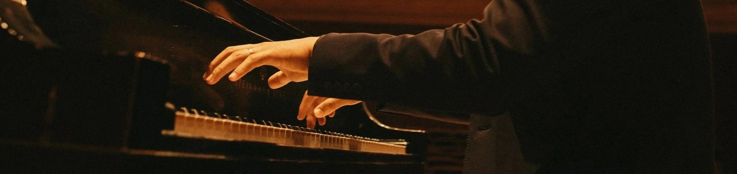 Person playing piano, wearing a dark jacket, with focus on hands on keyboard.