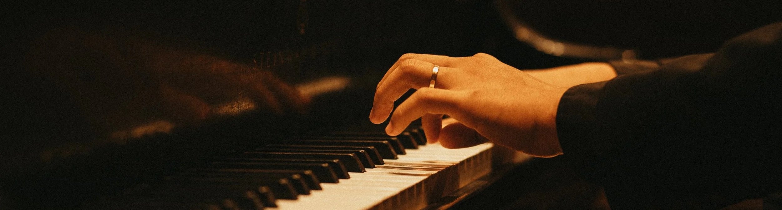 A person playing the piano, with a focus on their hand on the keys, wearing a ring on their finger in a dimly lit setting.