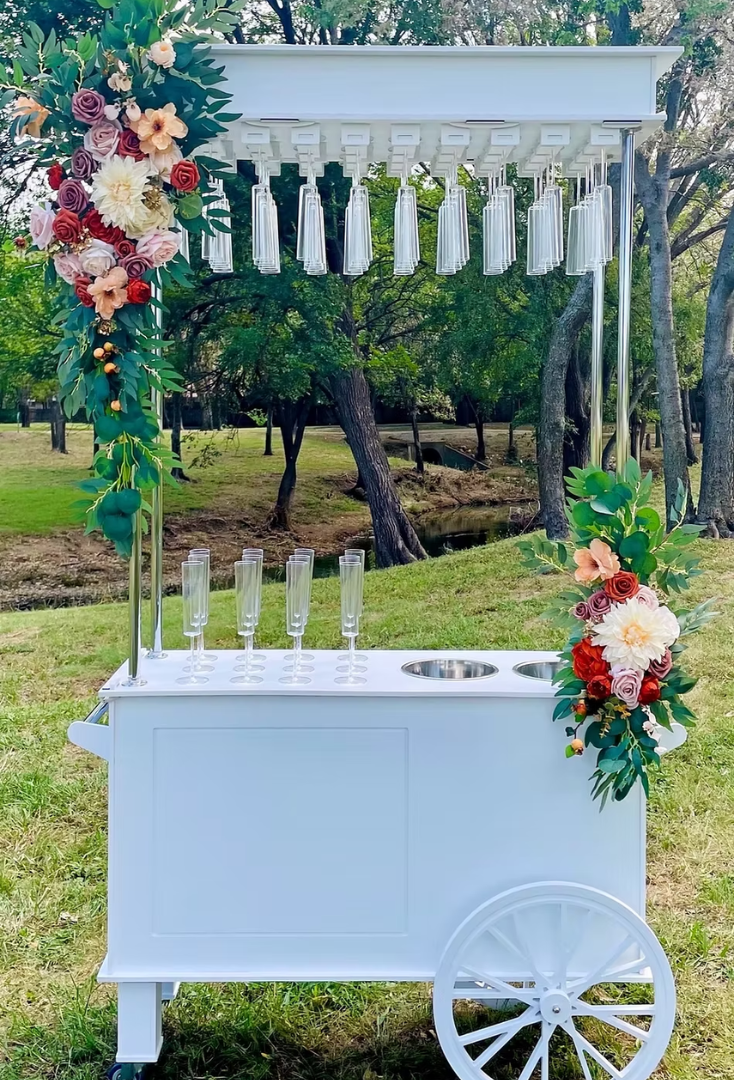 A white outdoor drink cart decorated with colorful flowers, featuring hanging glass cups and four empty champagne flutes, set in a park with trees in the background.