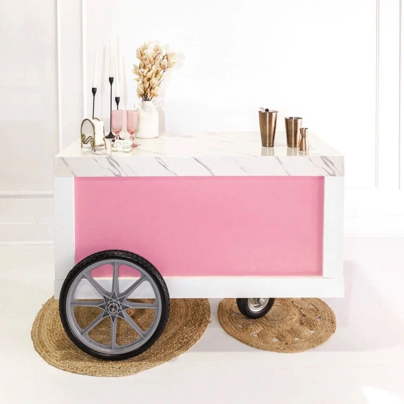 The Rose mobile bar rental by Sipsy Schooner pink bar for weddings and bridal showers in OKC holders, and decorative items including vases and candles, partially sitting on woven placemats with wheels.