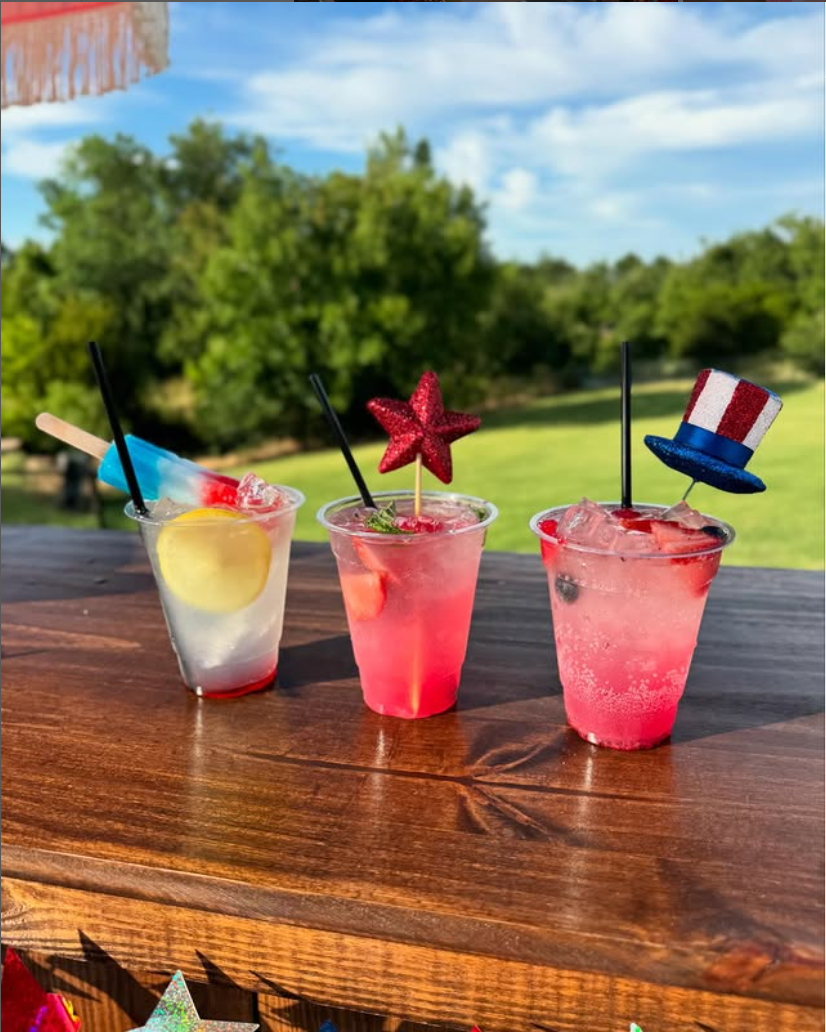 Three patriotic-themed drinks on a wooden table outdoors with trees and sky in the background. The first drink has a popsicle and lemon slice, the second has a red star decoration, and the third has an Uncle Sam hat decoration.