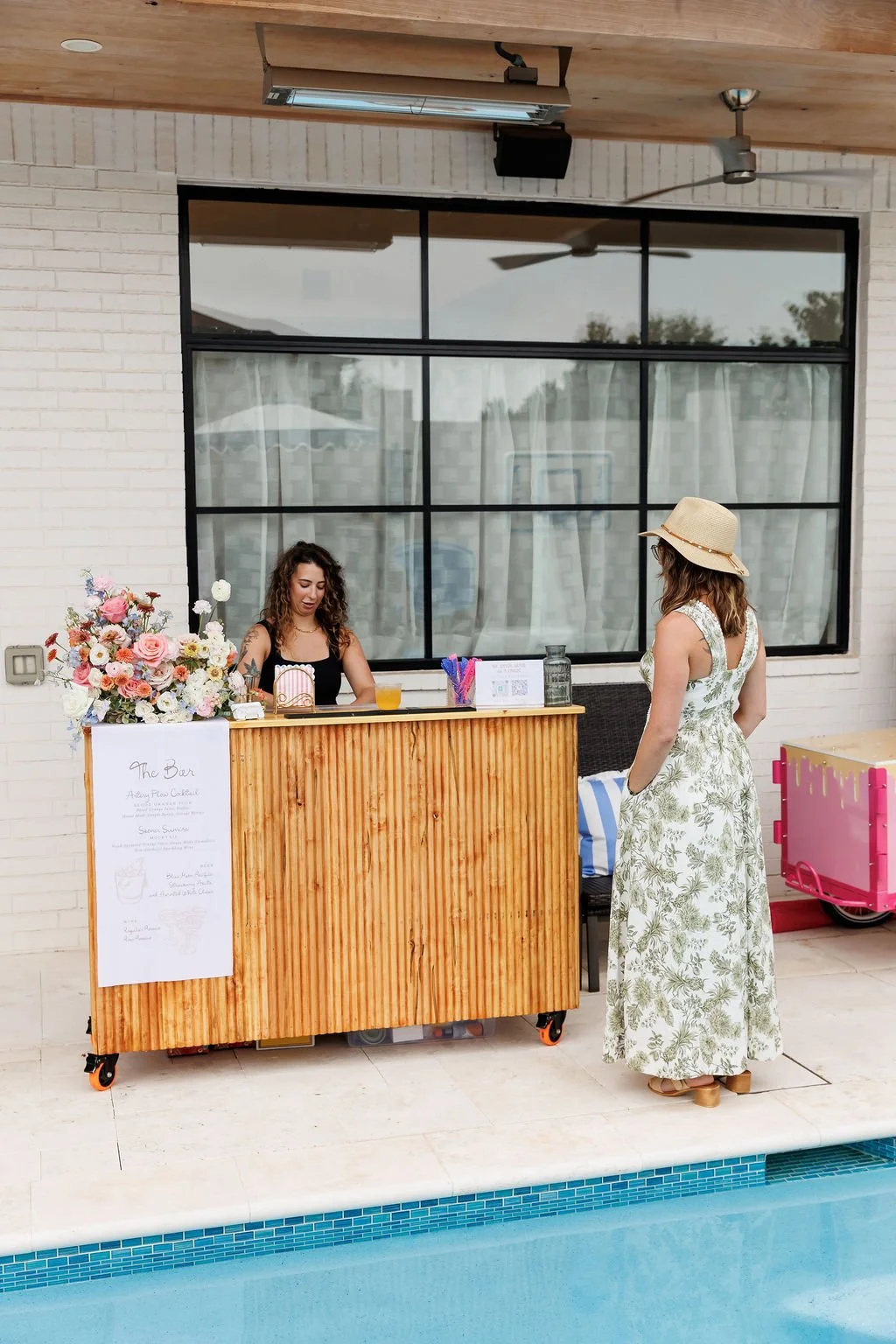A woman wearing a wide-brimmed hat and a floral maxi dress standing by a poolside bar with a bartender preparing a drink, decorated with a large bouquet of flowers, against a white brick wall and large window.
