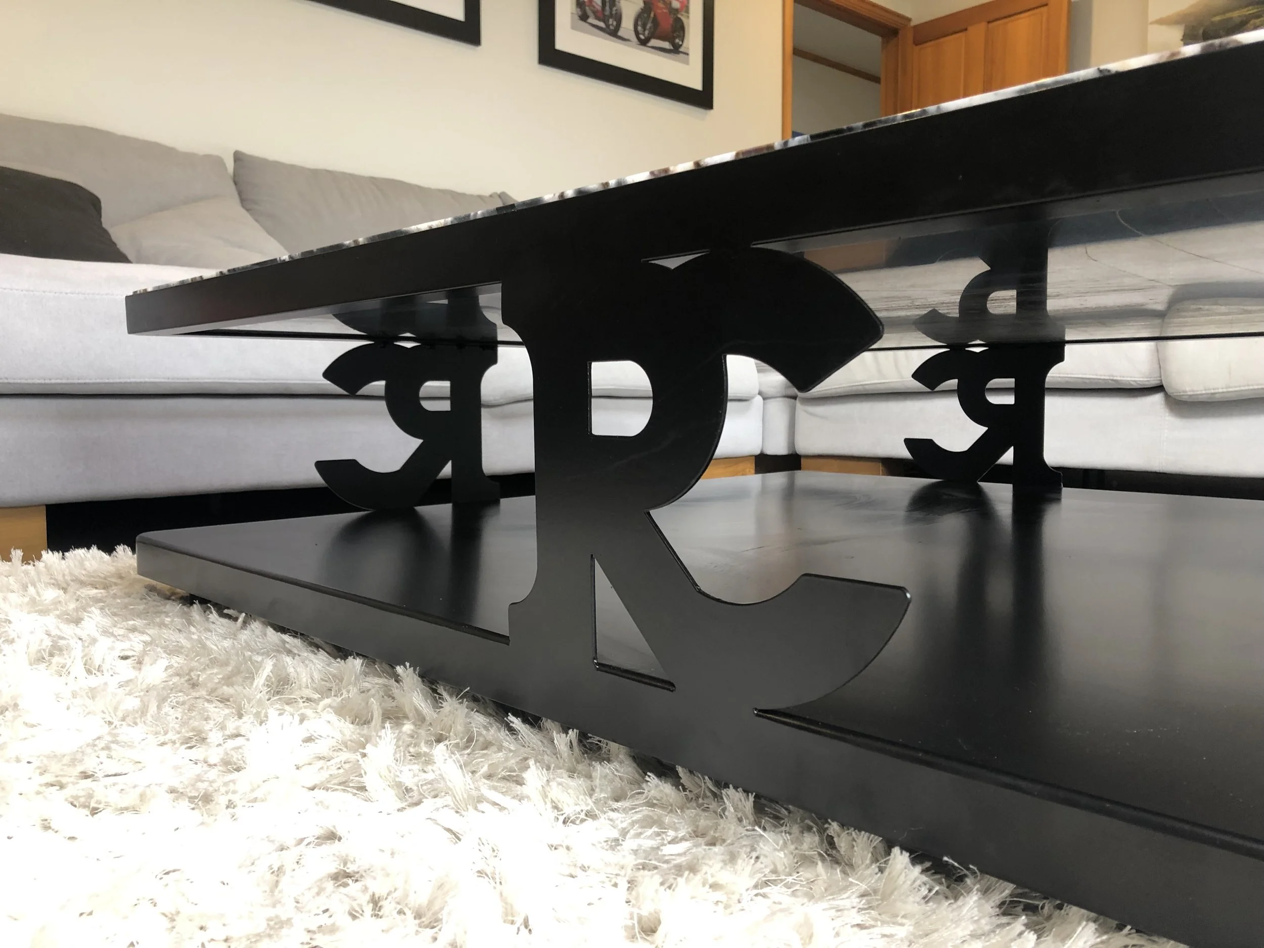 Black coffee table with decorative metal letter "R" supports, placed on a light shaggy rug in a living room with gray sectional sofa.