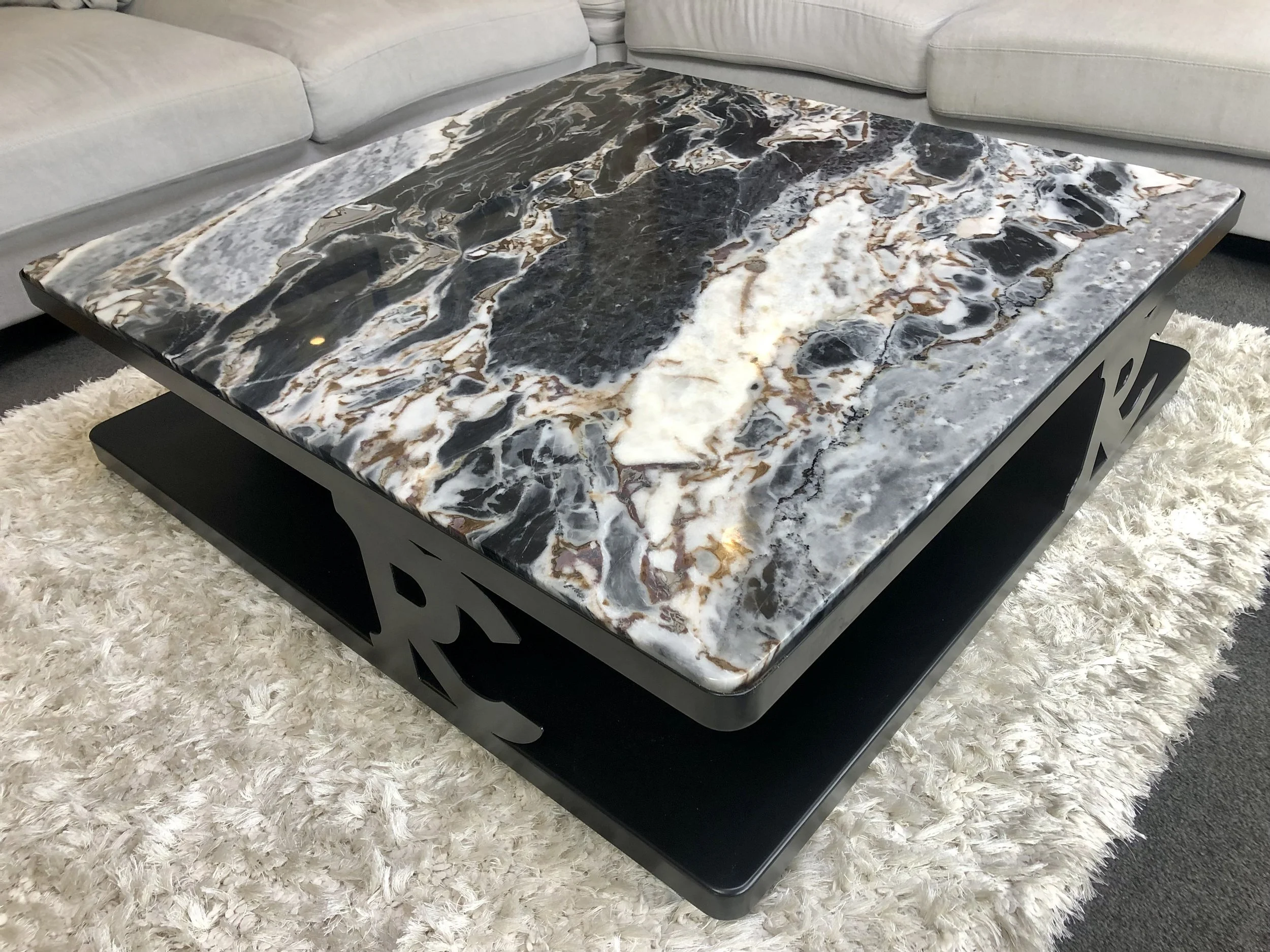 Modern square coffee table with a black and white marble top, featuring artistic metal side panels and a lower black shelf, resting on a fluffy white rug in a living room with a light-colored sofa.