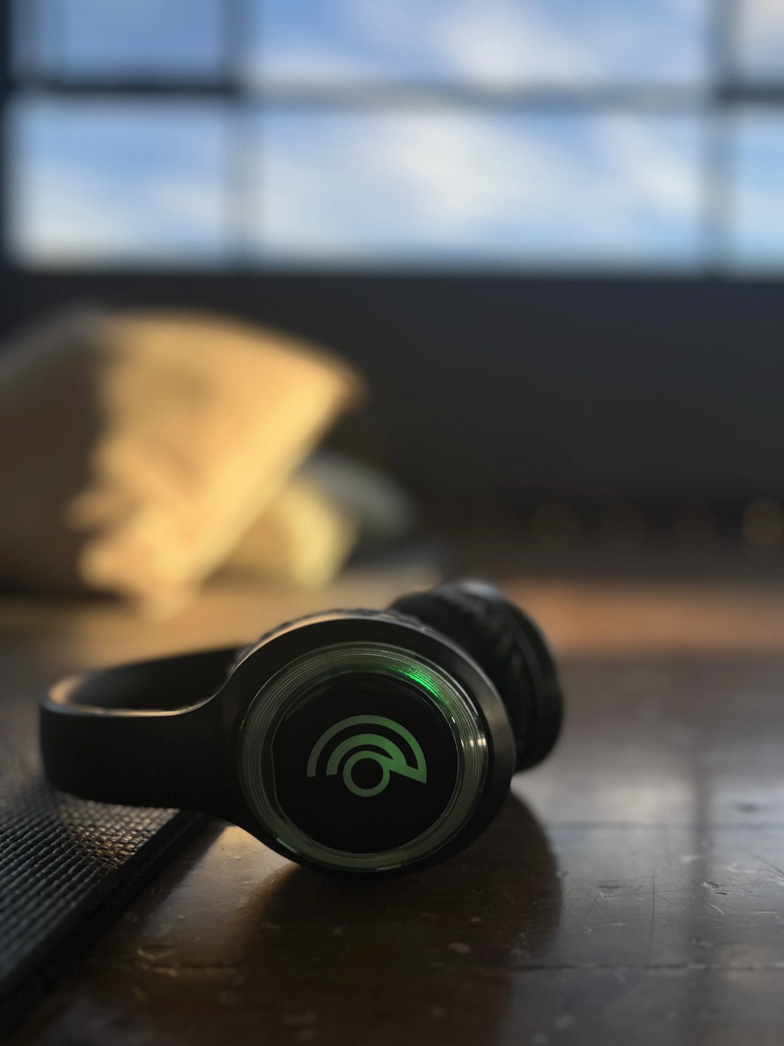 Close-up of black wireless earbuds with green indicators, resting on a wooden surface with a blurred background of a window with blue sky and a yellow object.