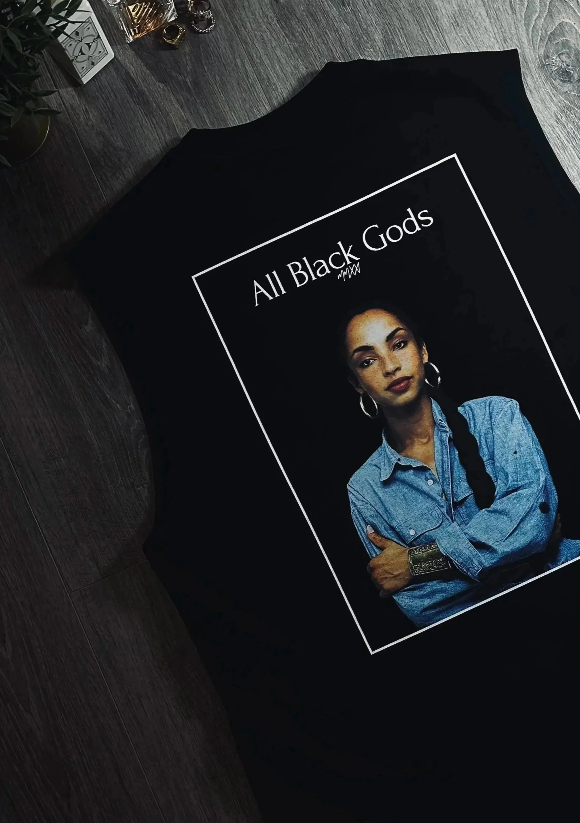 A black T-shirt with a photo of a woman with braided hair, wearing a denim shirt and hoop earrings. The text on the T-shirt reads "All Black Gods".