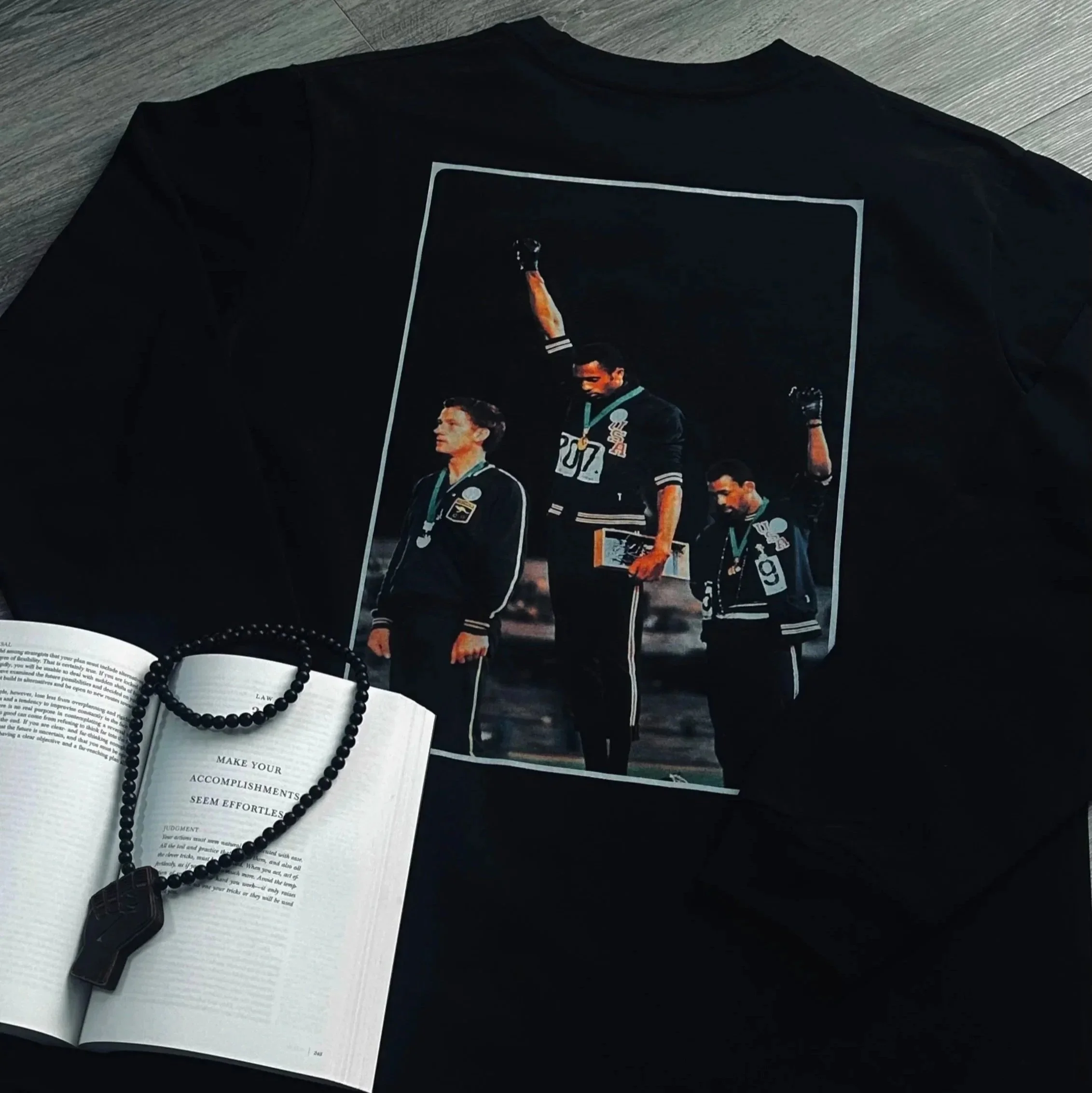 A black T-shirt featuring an image of three athletes standing on a podium with medals, one raising a fist, and two holding awards. An open book and a beaded necklace are in front of the shirt.