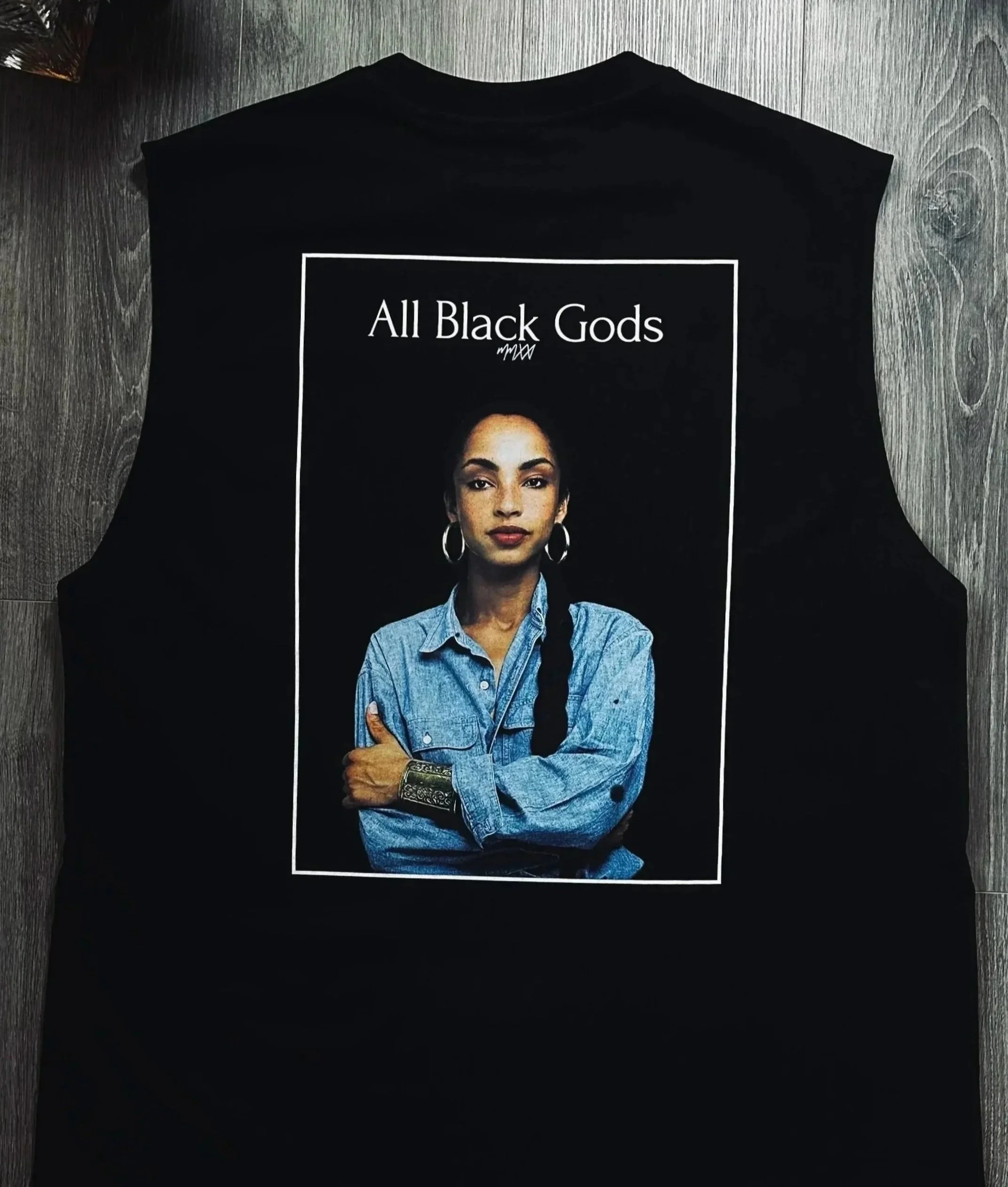 Black sleeveless T-shirt featuring a woman with dark hair, wearing hoop earrings and a denim shirt, with the text "All Black Gods" above her. The T-shirt is laid flat on a wooden surface.