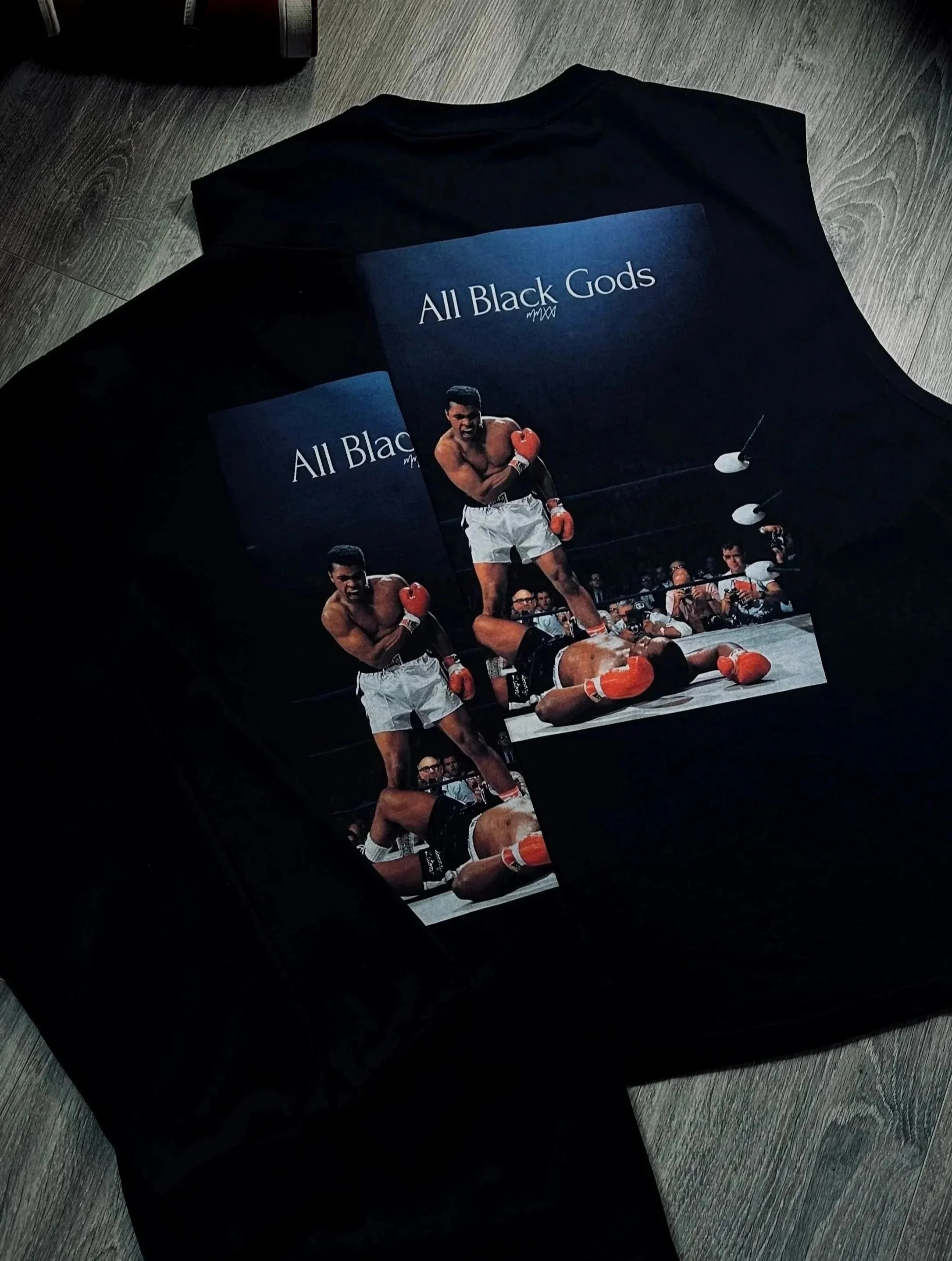 A black t-shirt with a printed photograph of a boxing match, showing two fighters in the ring, one standing over the other who is on the ground, with the text "All Black Gods" above the image.