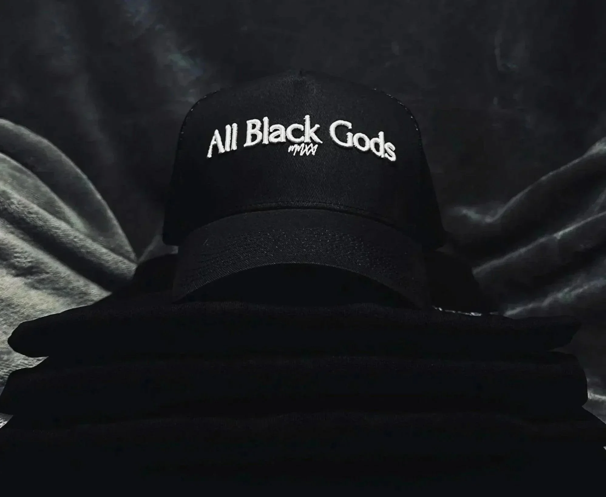 Black baseball cap with embroidered white text that reads 'All Black Gods' placed on folded black clothing against a dark, textured background.