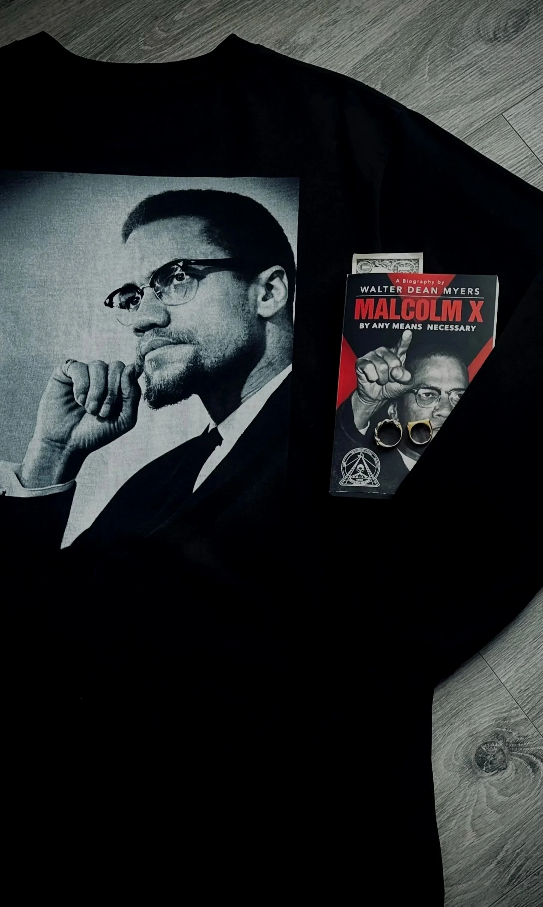 A black t-shirt featuring a large grayscale portrait of Malcolm X, with a book about Malcolm X and some rings placed on it. The book has a red and black cover with an image of Malcolm X and some symbols.