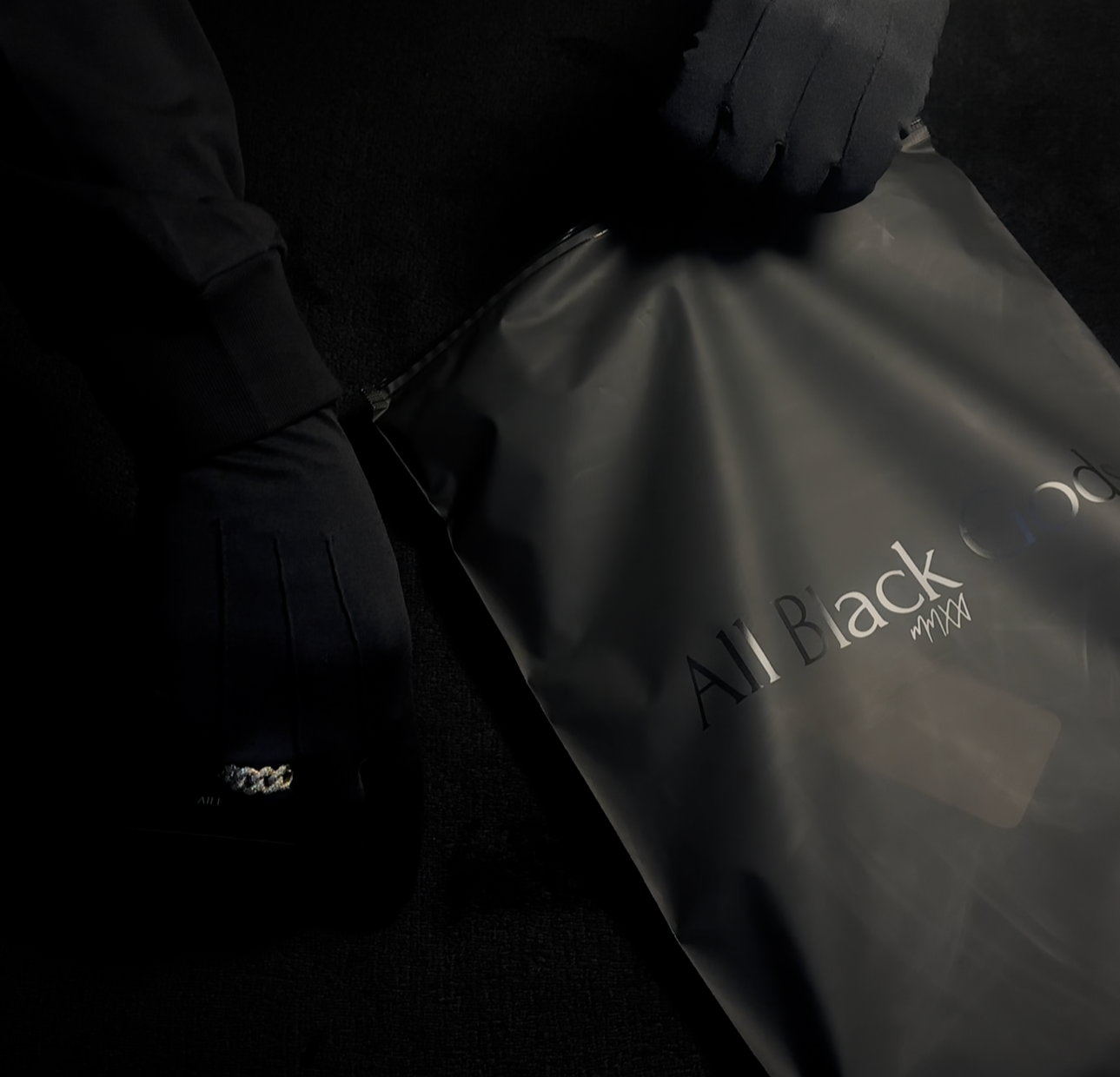 A person wearing black gloves and black pants, sitting with their legs crossed on a dark surface. Next to them is a black shopping bag with the text 'All Black' printed on it.
