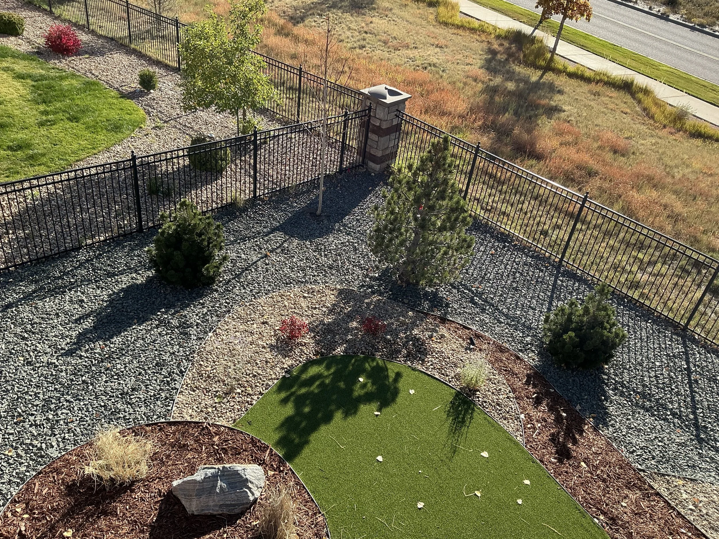 A landscaped backyard with a black metal fence, small trees, bushes, and rocks, along with a green artificial turf area.