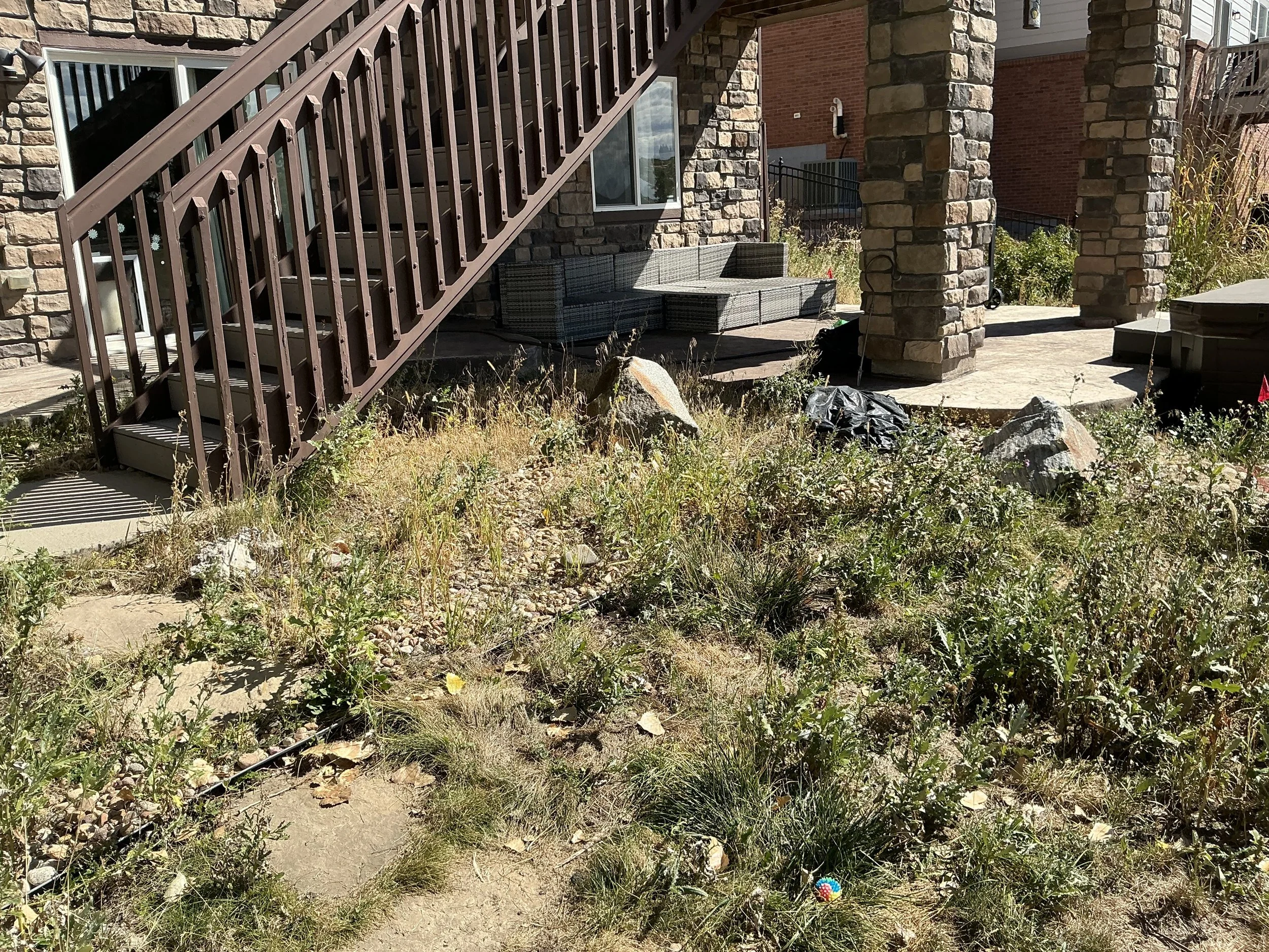 Backyard patio area with stone columns, outdoor seating, and a staircase leading to the upper level, surrounded by a garden with weeds and rocks.