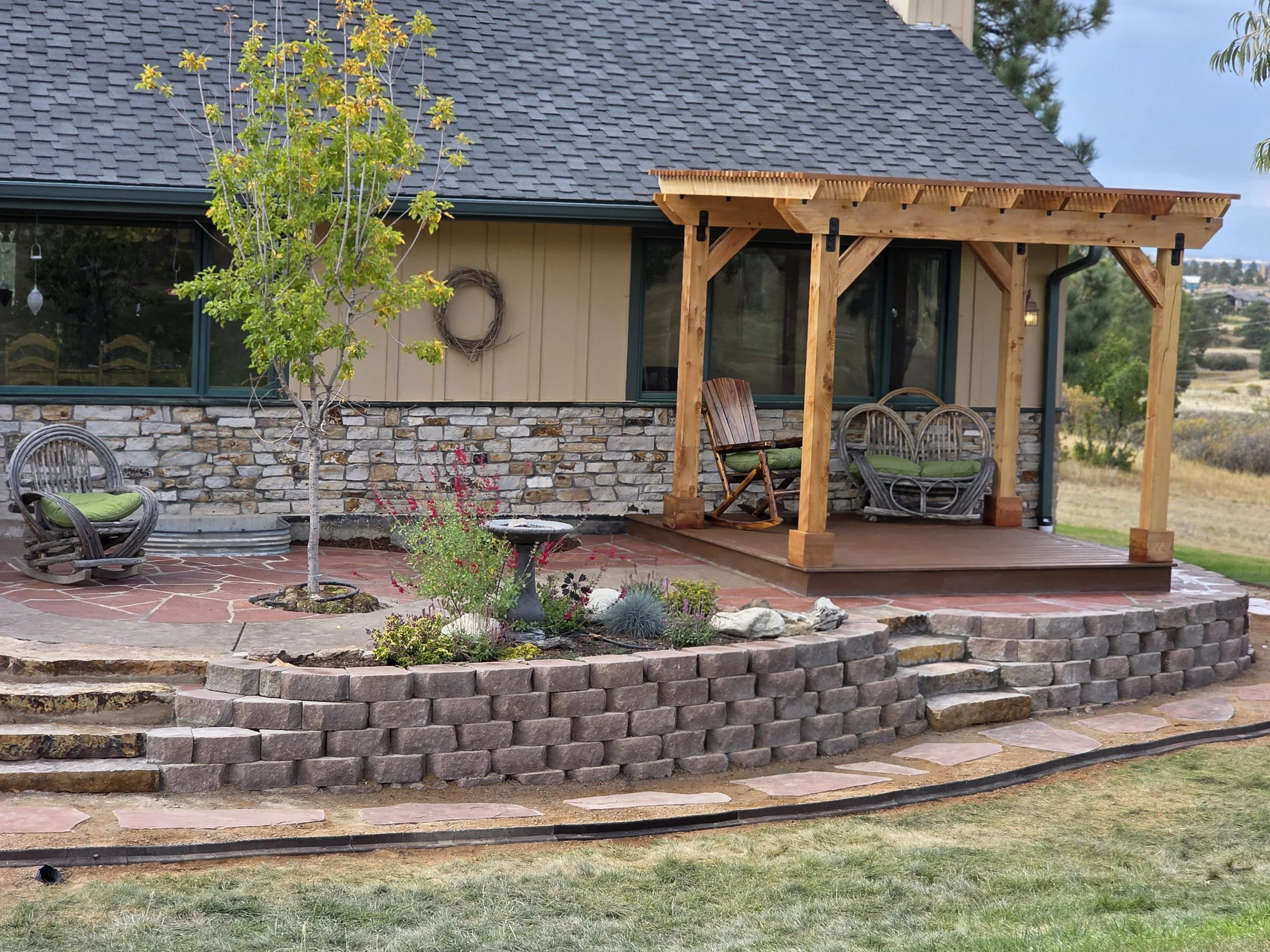 A backyard patio with a small garden, three wicker chairs with green cushions, one wooden rocking chair on a wooden deck under a pergola, a small tree, and a stone wall with a window and decorative wreath.