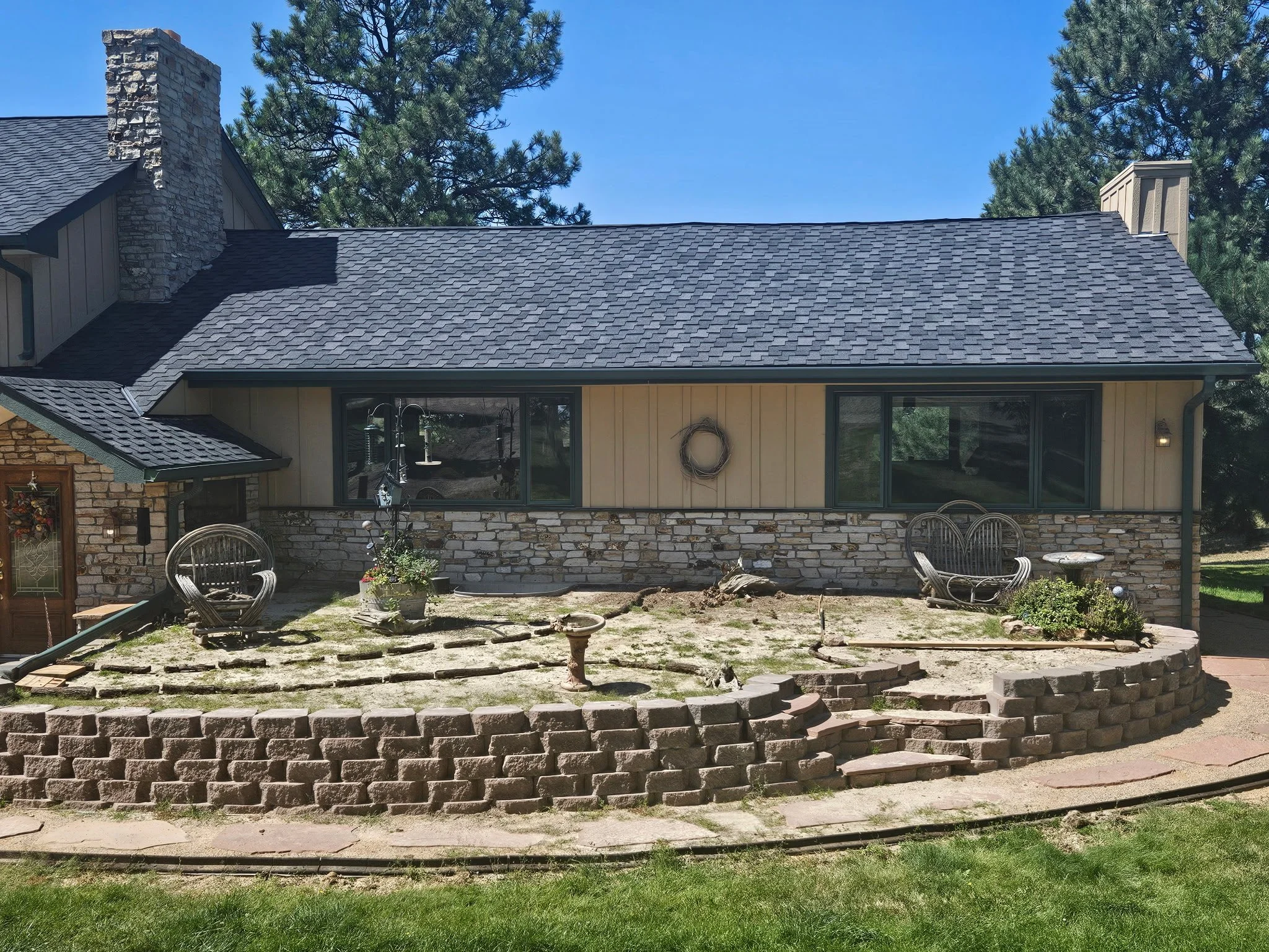Front view of a house with a stone and beige siding exterior, dark gray shingle roof, large windows, and a small garden area with outdoor seating, a birdbath, and some plants.