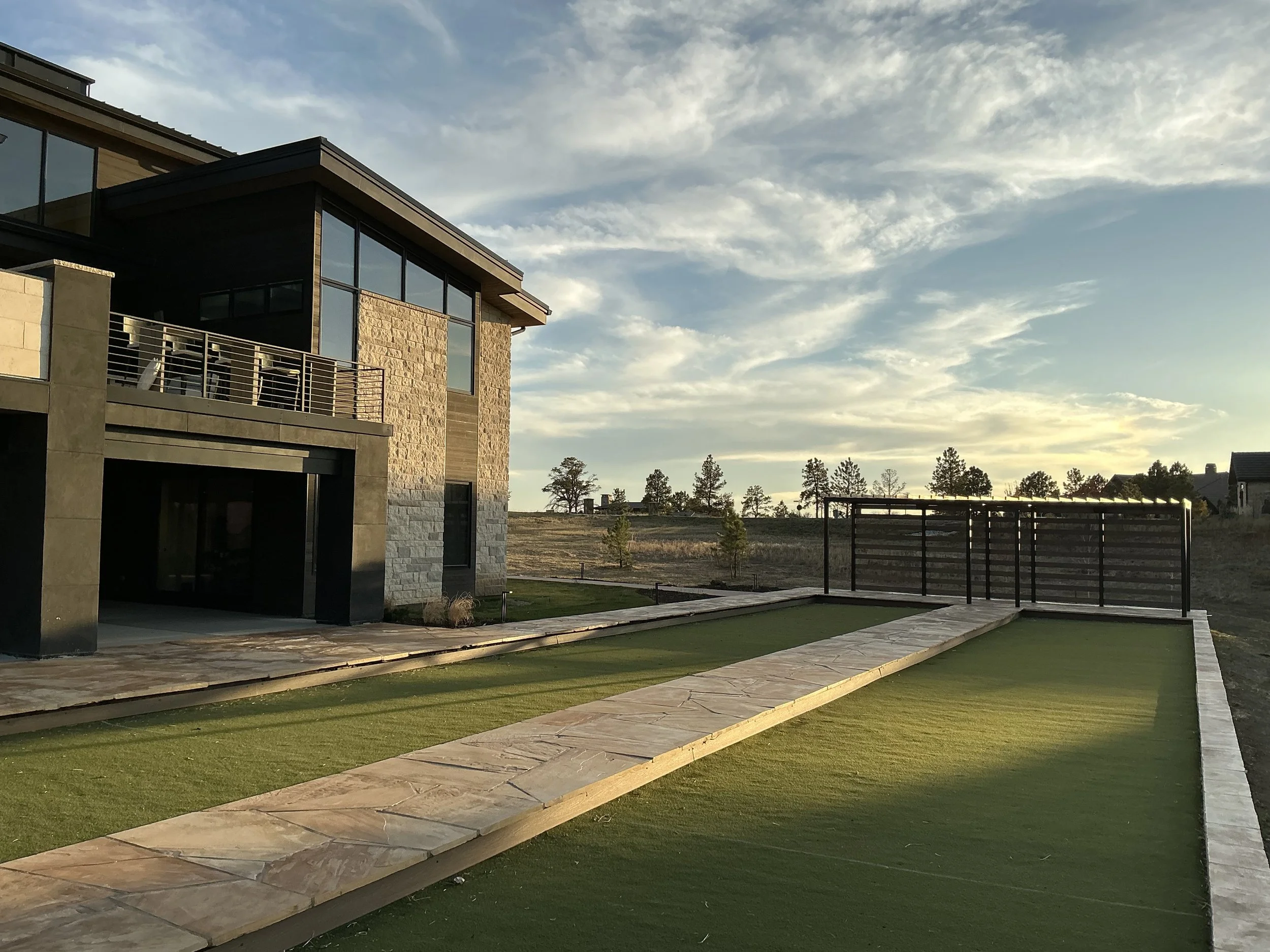 Modern house with large glass windows, balcony, and stone exterior, adjacent to a lawn and outdoor shuffleboard court, under a partly cloudy sky at sunset.