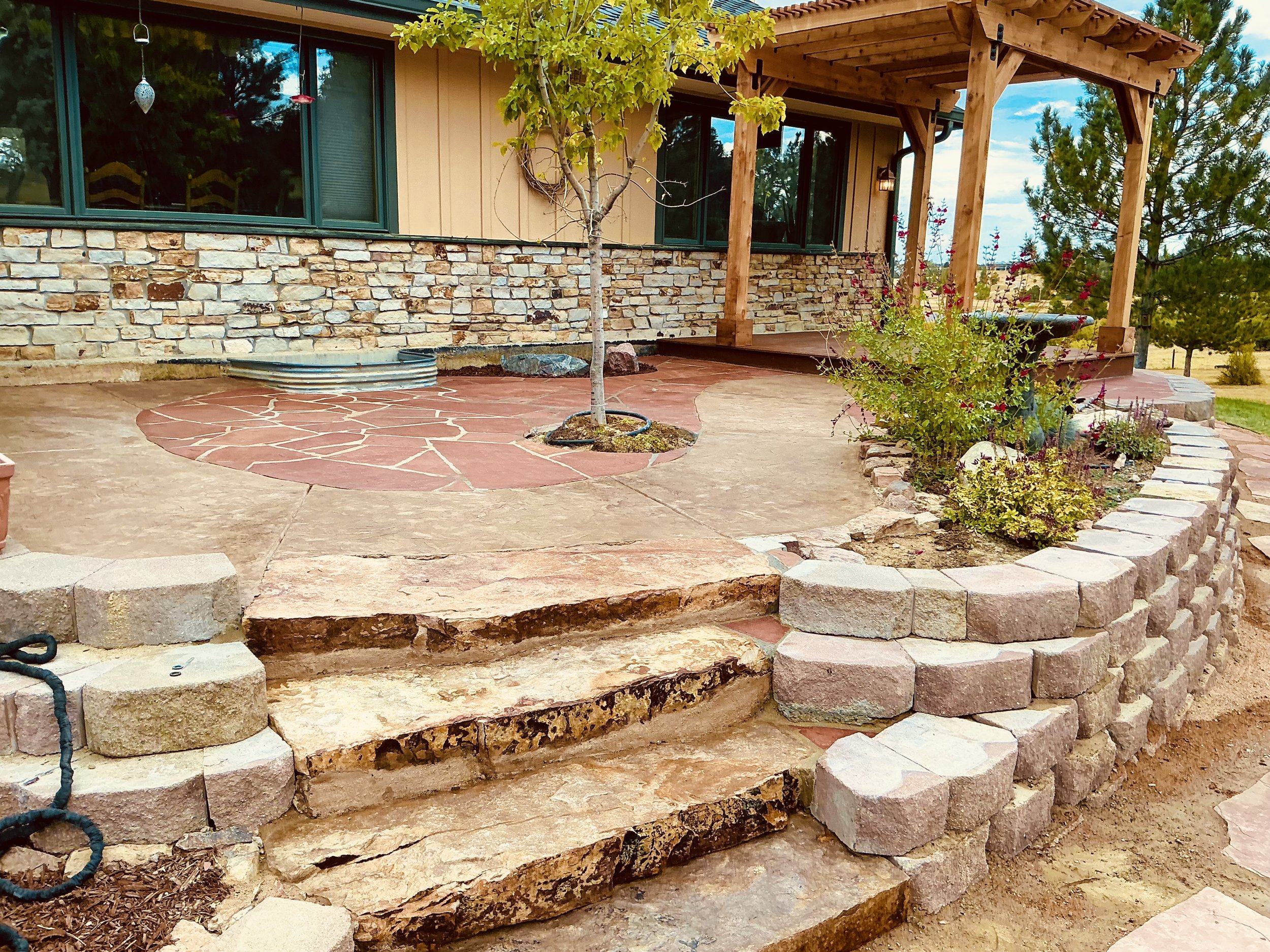 Backyard patio with stone stairs, a small tree, a wooden pergola, and a planter with flowers.