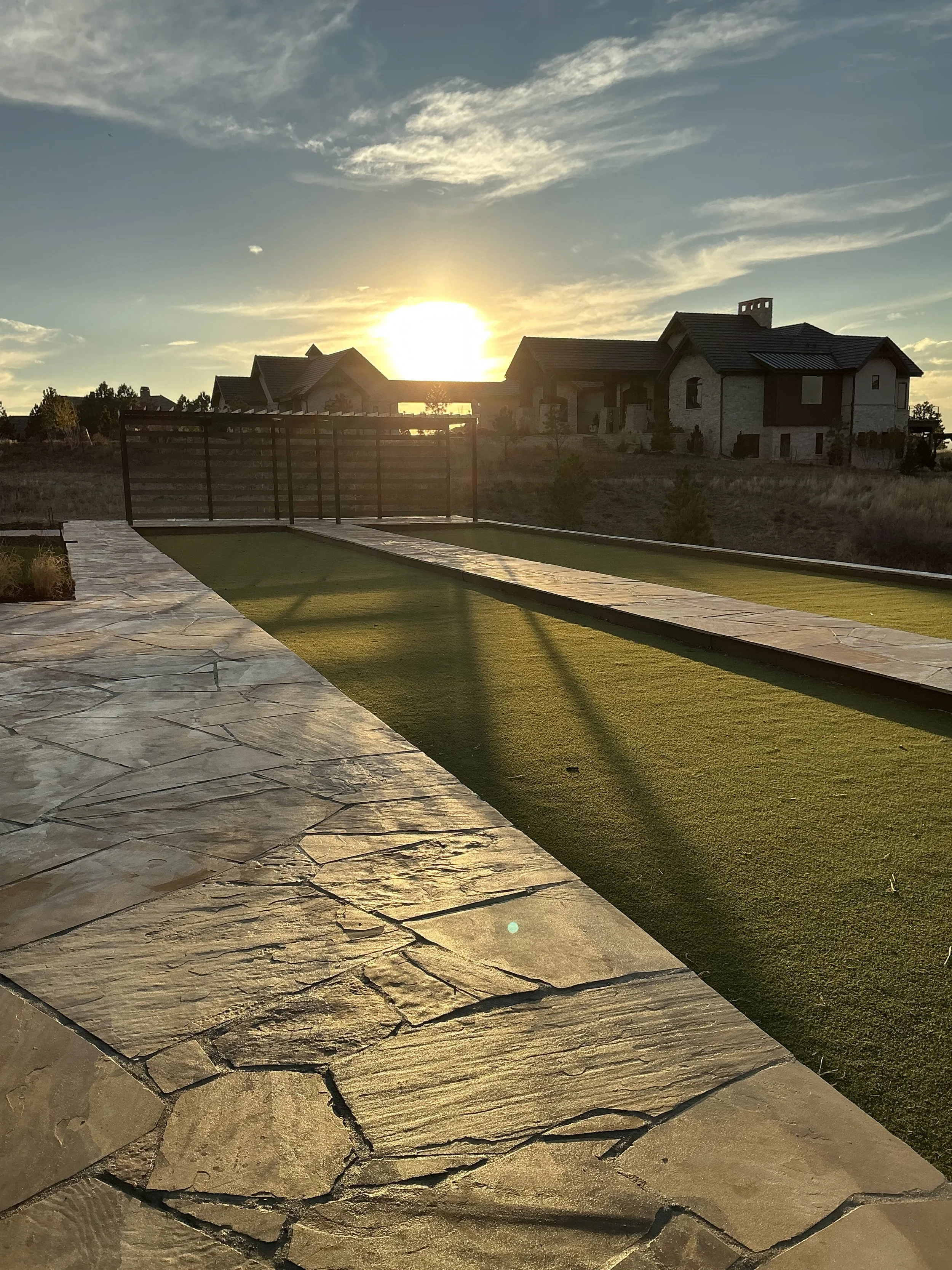 Sunset over a backyard with artificial turf and stone patio, with houses in the background.