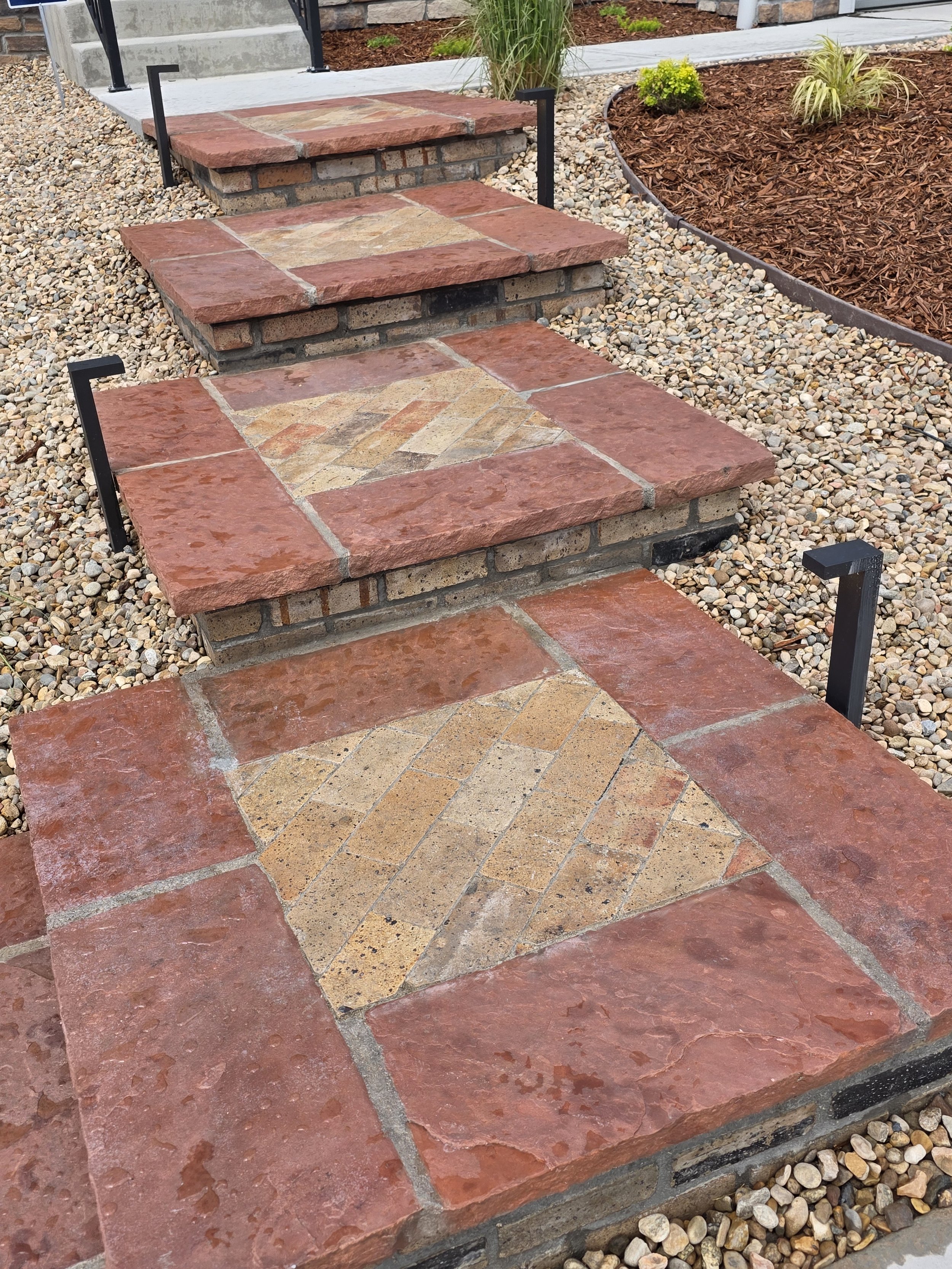 Outdoor brick steps with metal handrails leading up to a porch, surrounded by gravel and landscaped flower beds.