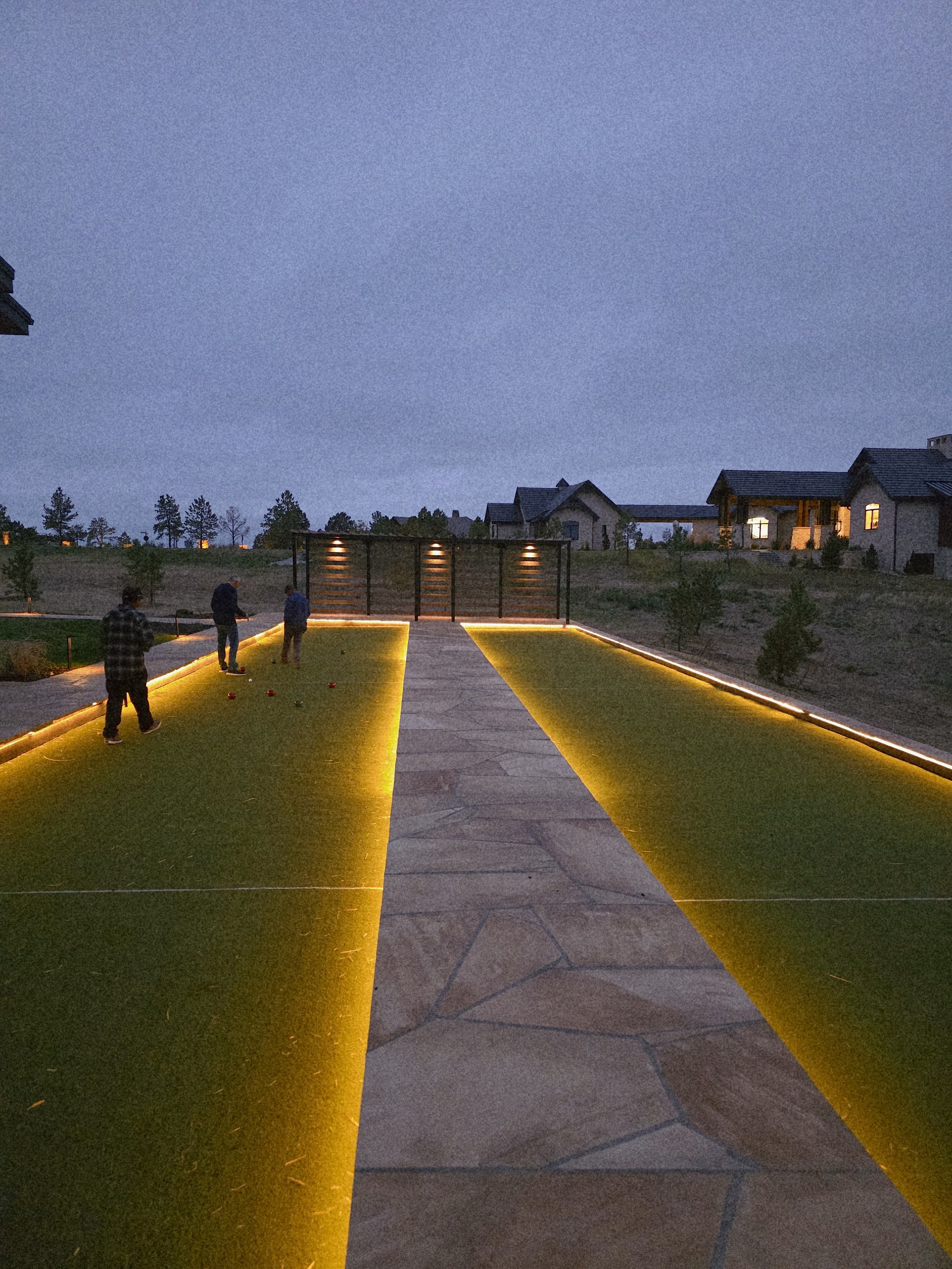 People playing bocce ball on a modern outdoor bocce court during dusk, with LED lighting along the edges.