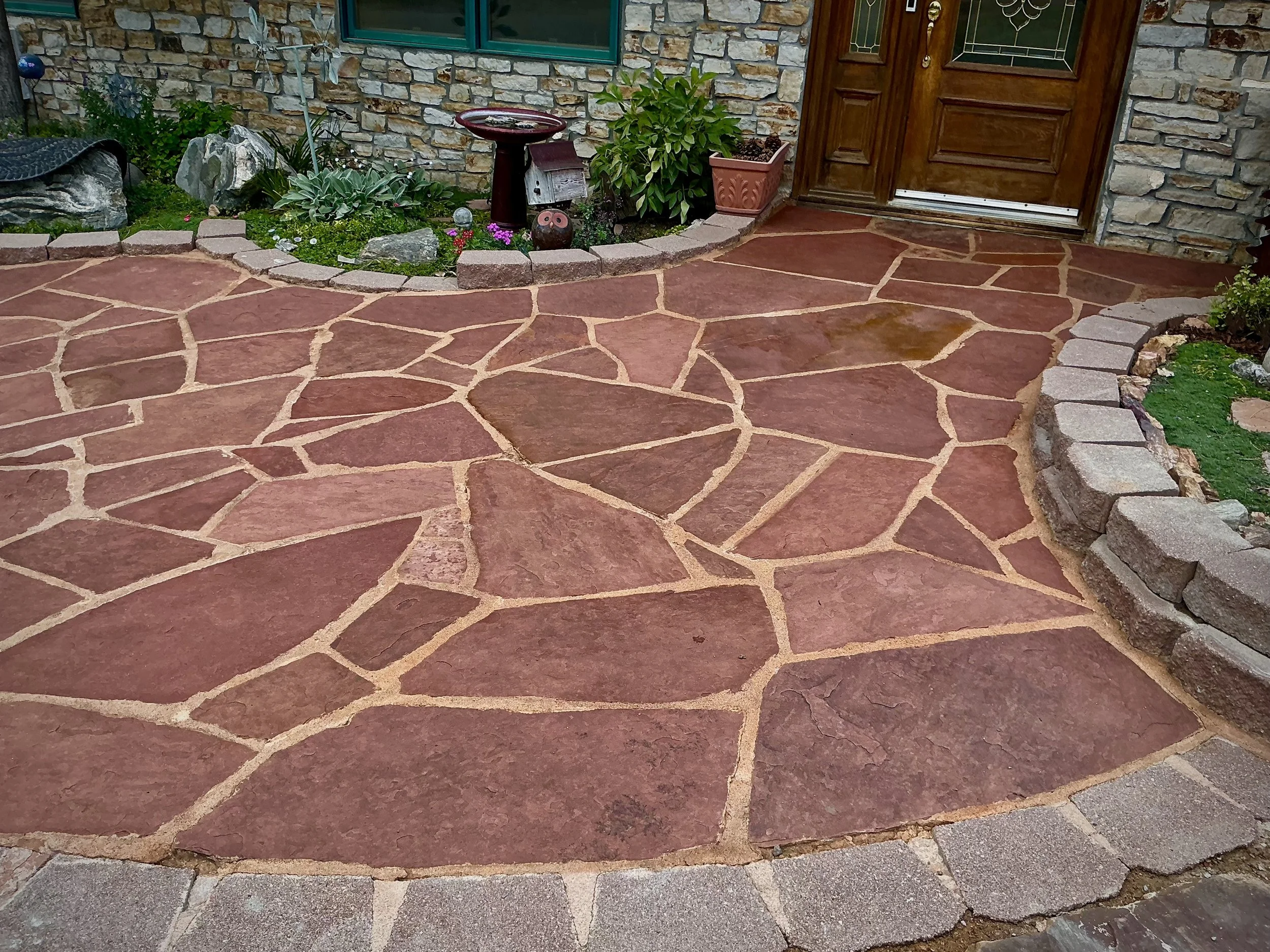 Red flagstone pathway with a stone border leading to a wooden front door of a house, with plants and garden decor along the sides.