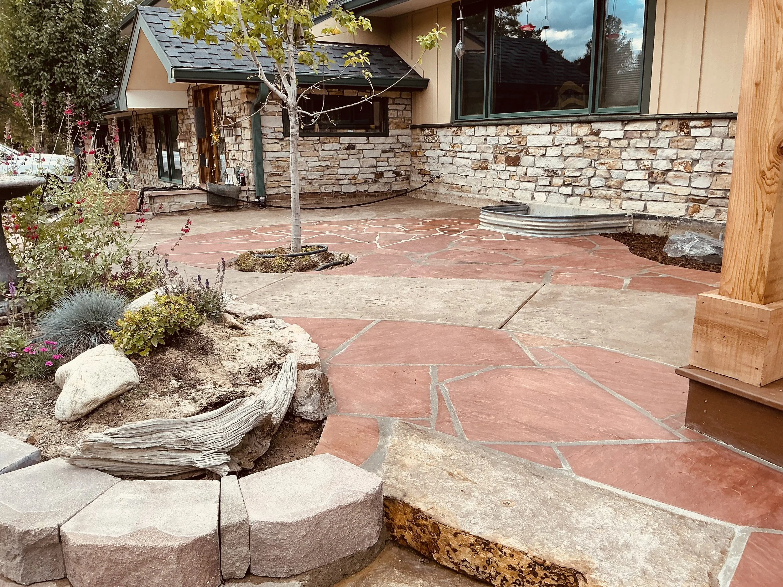 Photo of a backyard patio with a stone house, leafless tree, potted plants, and a combination of large irregular red and beige paving stones, with a small garden bed on the left.