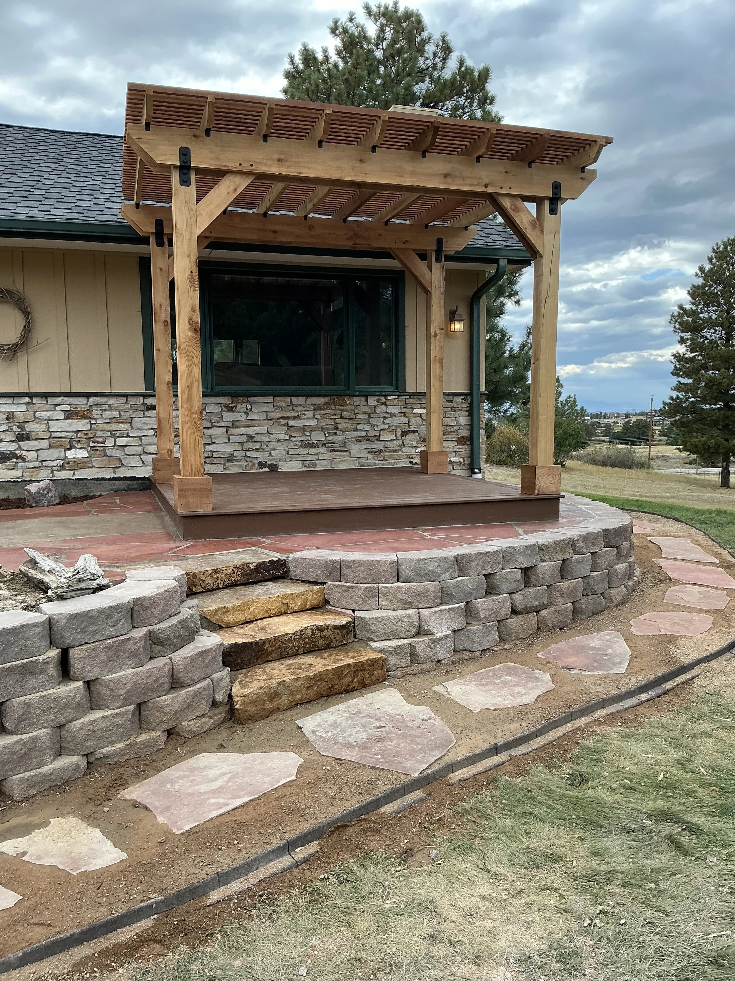 A newly built wooden pergola over a small porch area with stone and brick steps leading up to it, set against a house with stone and siding exterior and a large window. A landscaped pathway of flagstone surrounds the steps, and the yard has grass and trees in the background under a partly cloudy sky.