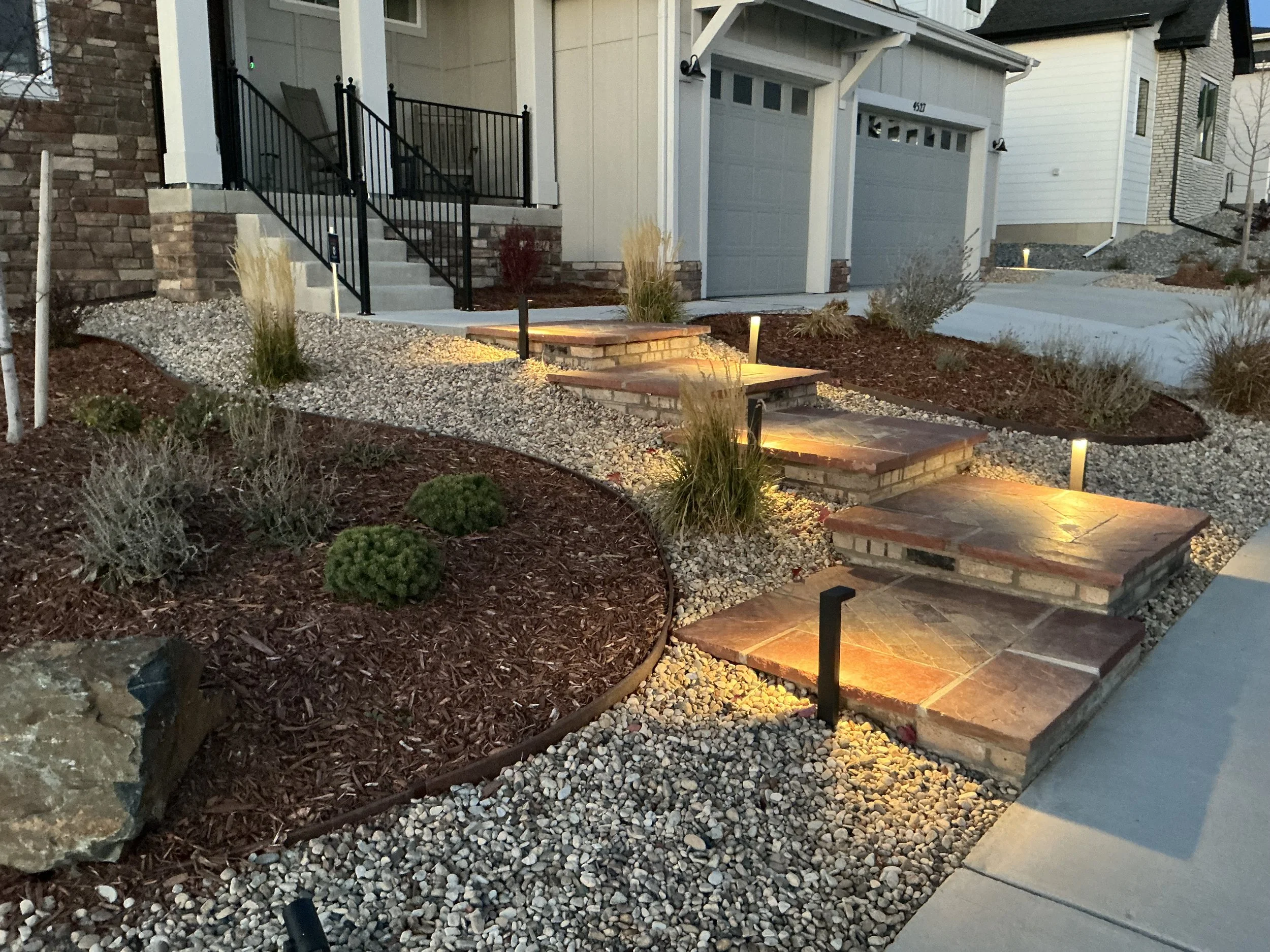 Exterior view of a modern house with a landscaped front yard, illuminated pathway lights, stone steps, and a concrete driveway.