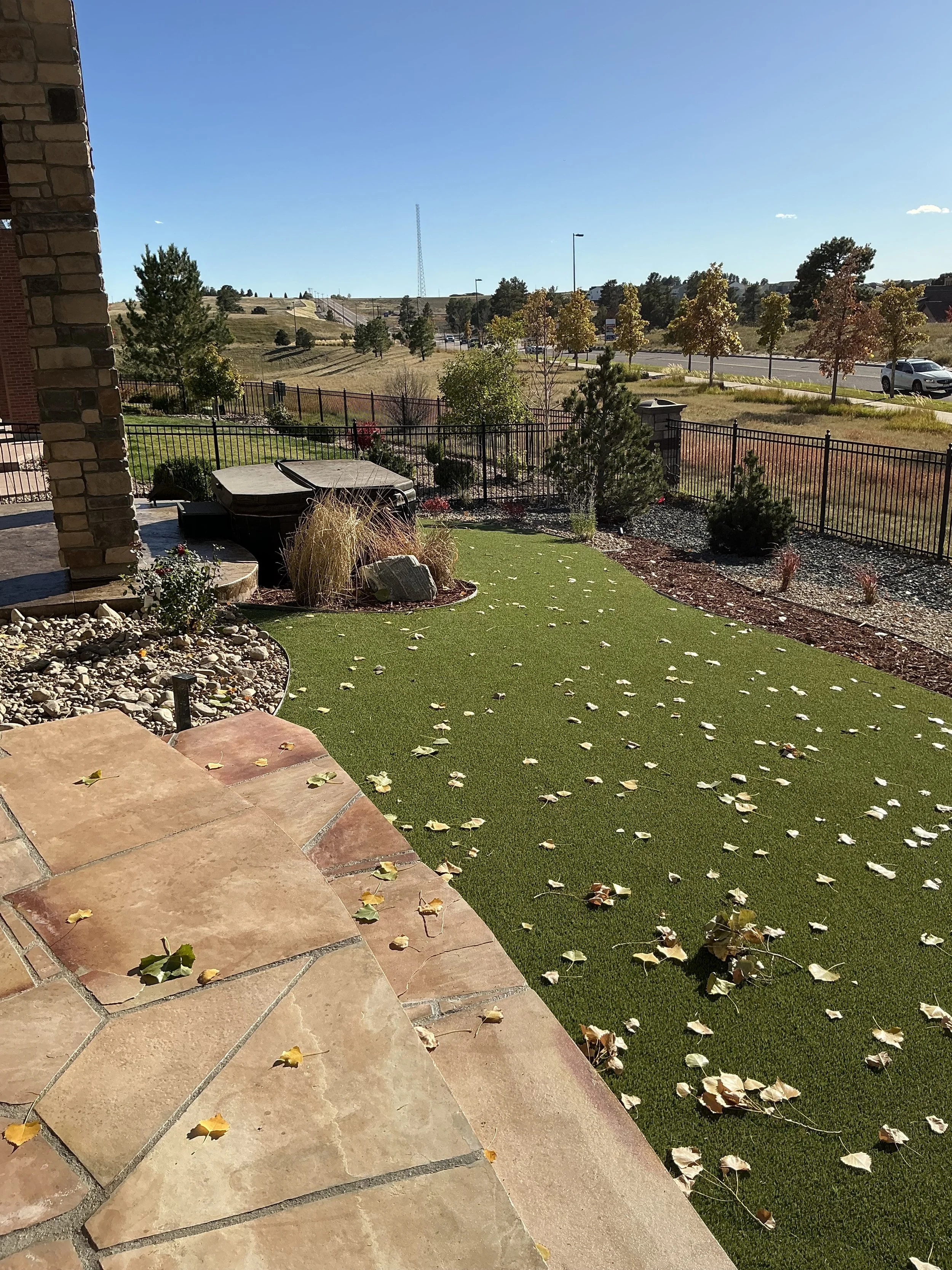Residential backyard with a stone patio, artificial grass with fallen leaves, trees, shrubs, garden bed with plants, and a black metal fence, with a view of a road and grassy area beyond.