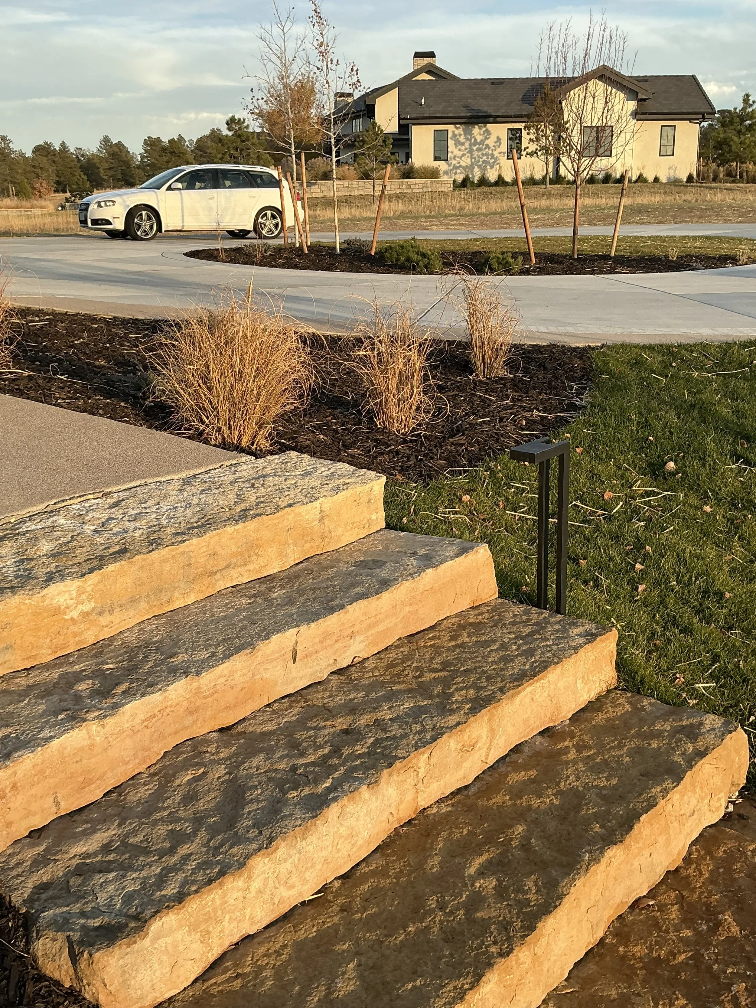 Stone steps leading up to a lawn with ornamental plants, in front of a house with beige siding and dark roof, in suburban neighborhood, during late afternoon.
